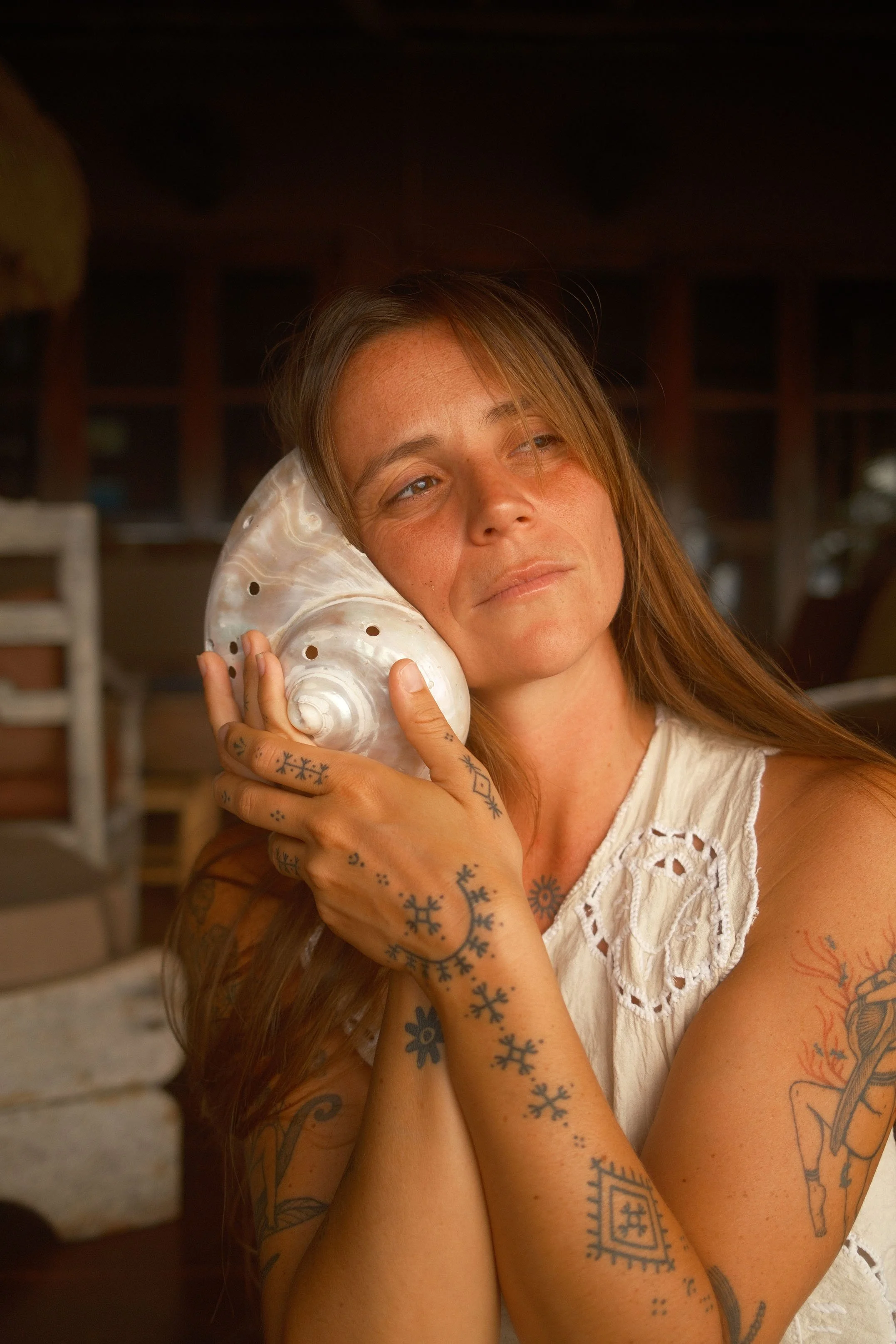 Femme avec tatouages portant un coquillage géant sur l'oreille, debout dans un environnement intérieur chaleureux.
