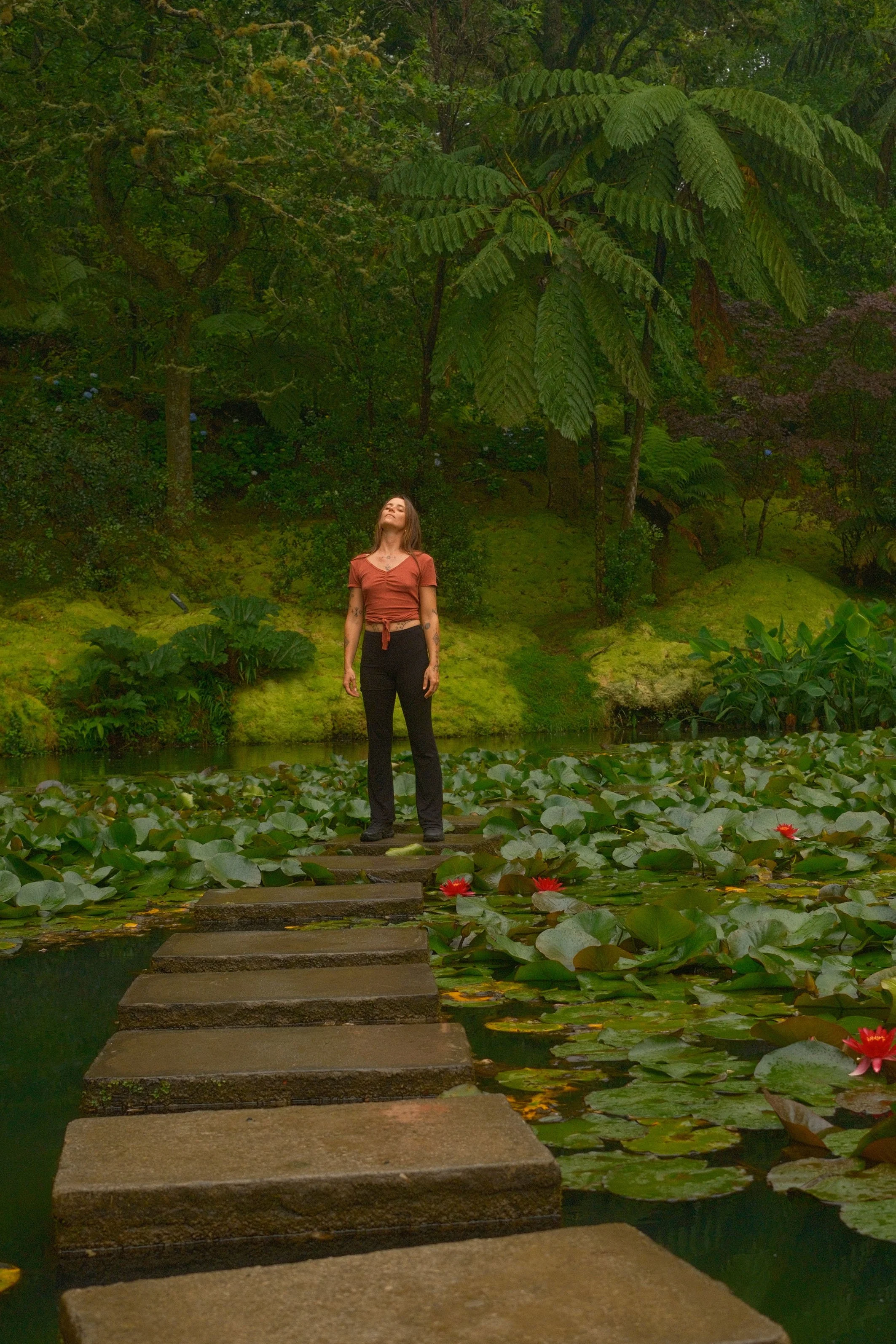 Une femme debout sur un petit pont en pierre au-dessus d'un étang rempli de nénuphars, entourée d'une végétation dense et de grands arbres dans une forêt ou un jardin tropical.