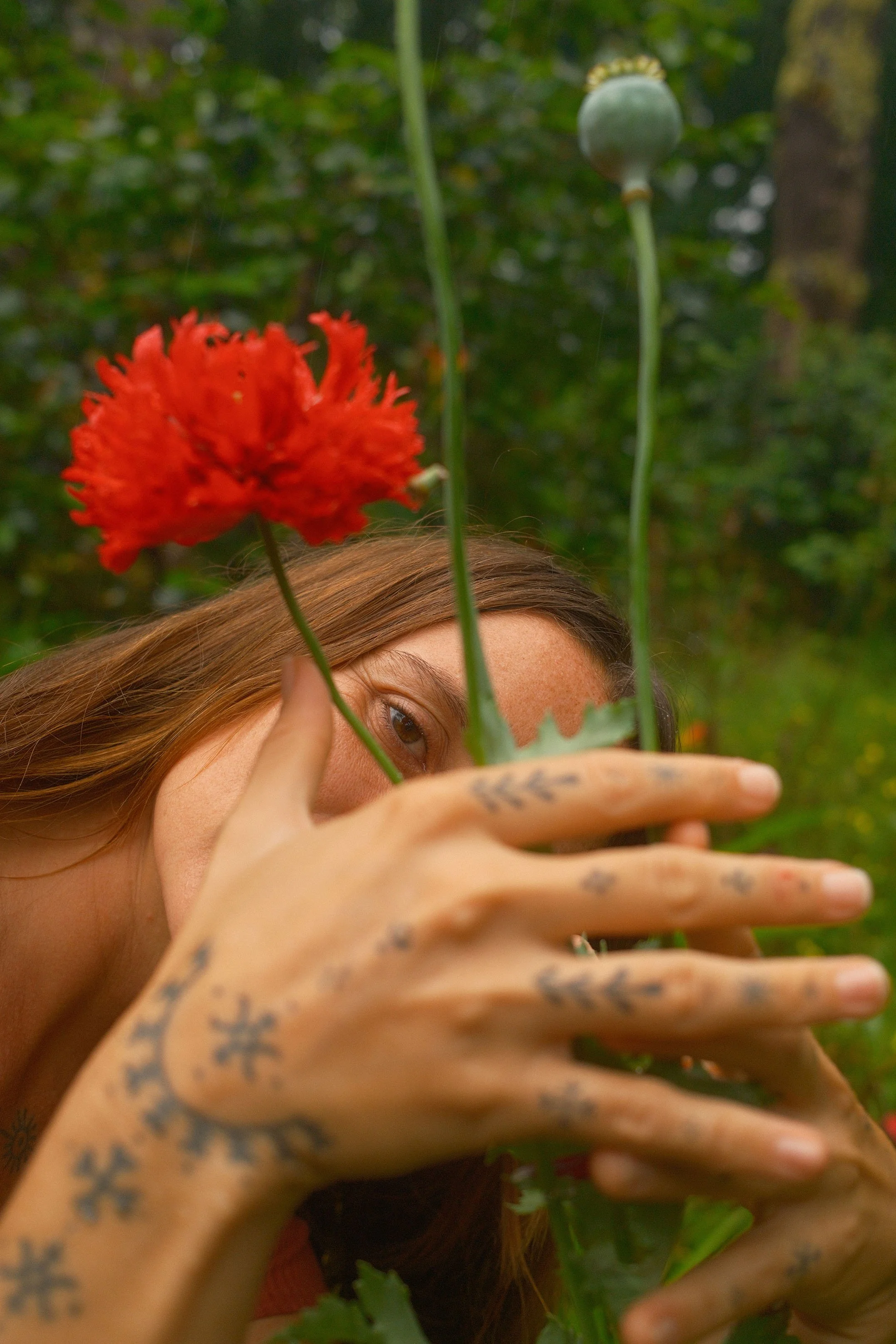 Une femme avec des tatouages sur les mains et des cheveux bruns, tenant une fleur rouge. Elle partiellement cachée derrière des tiges de fleurs et un fruit vert, dans un environnement naturel avec de la végétation en arrière-plan.