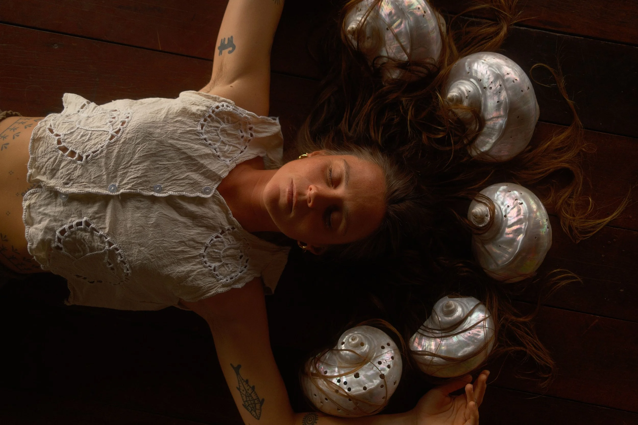 Femme allongée sur un sol en bois, entourée de coquillages en nacre, portant un vêtement blanc en dentelle, avec des tatouages sur le bras, les yeux fermés