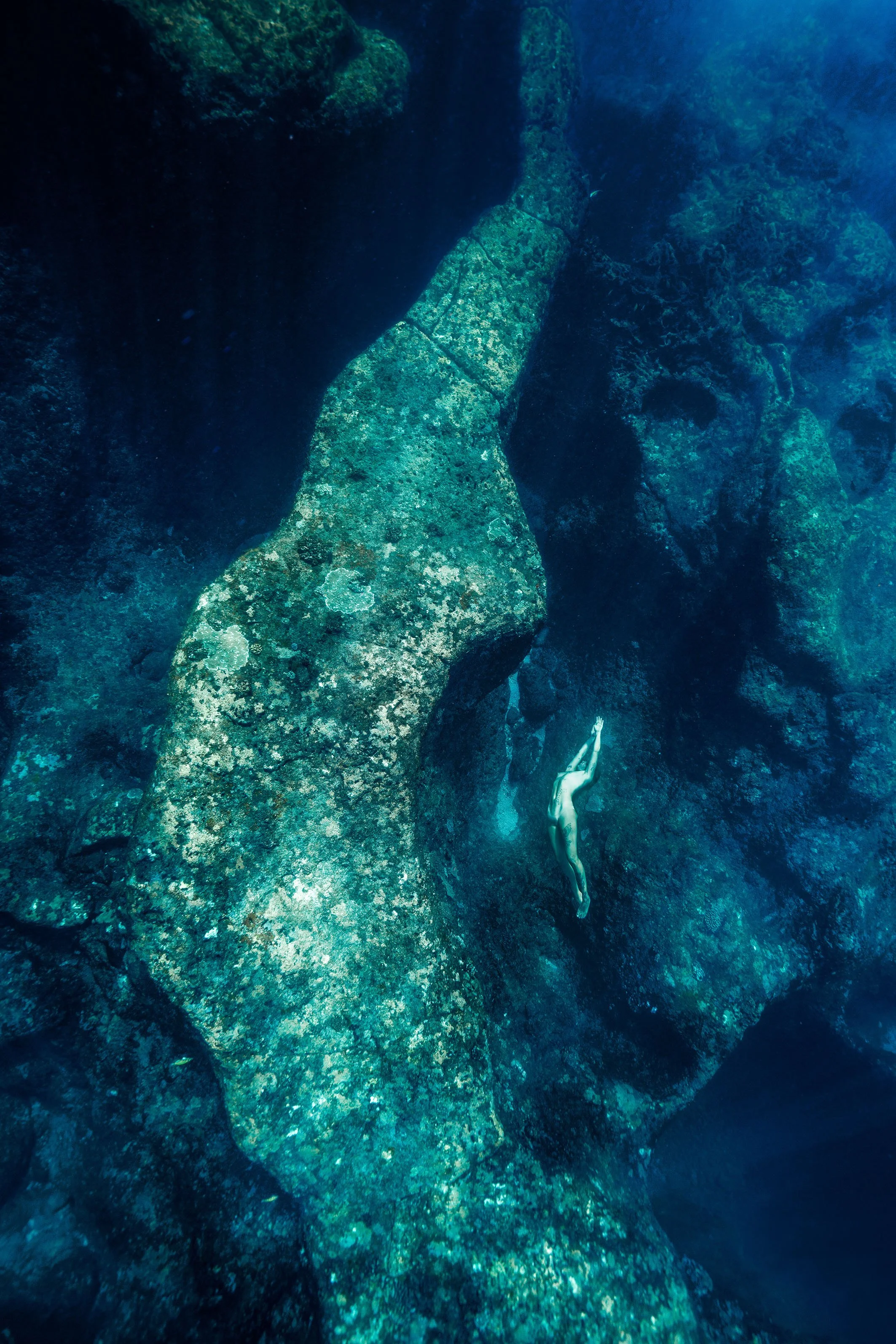 Une personne nue nage sous l'eau claire parmi des rochers et des formations rocheuses.