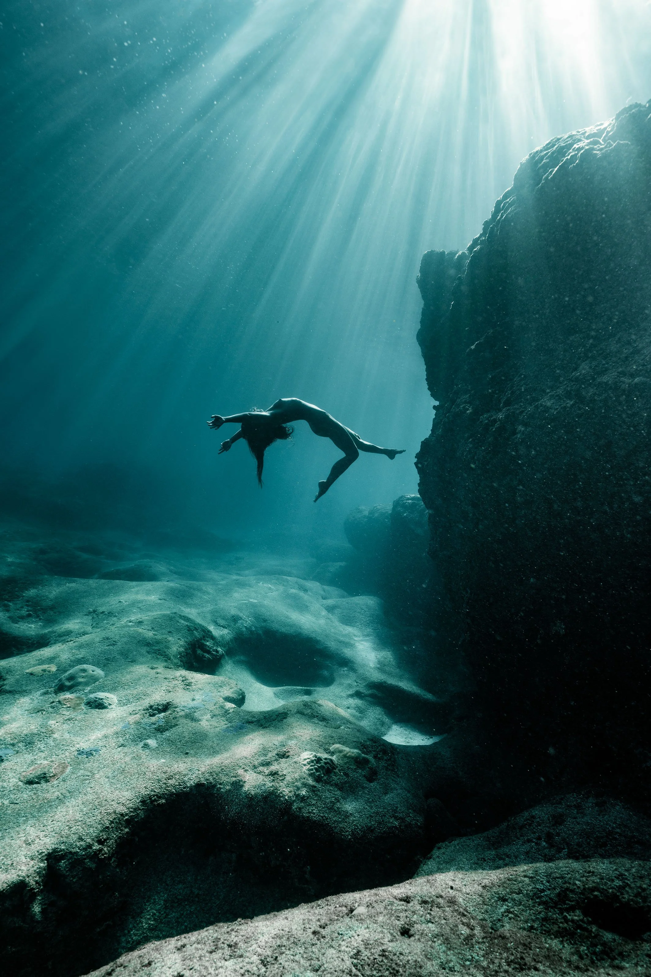 Une femme nage sous l'eau, éclairée par des rayons de soleil traversant la surface de l'eau, dans un environnement aquatique avec des rochers et du sable.