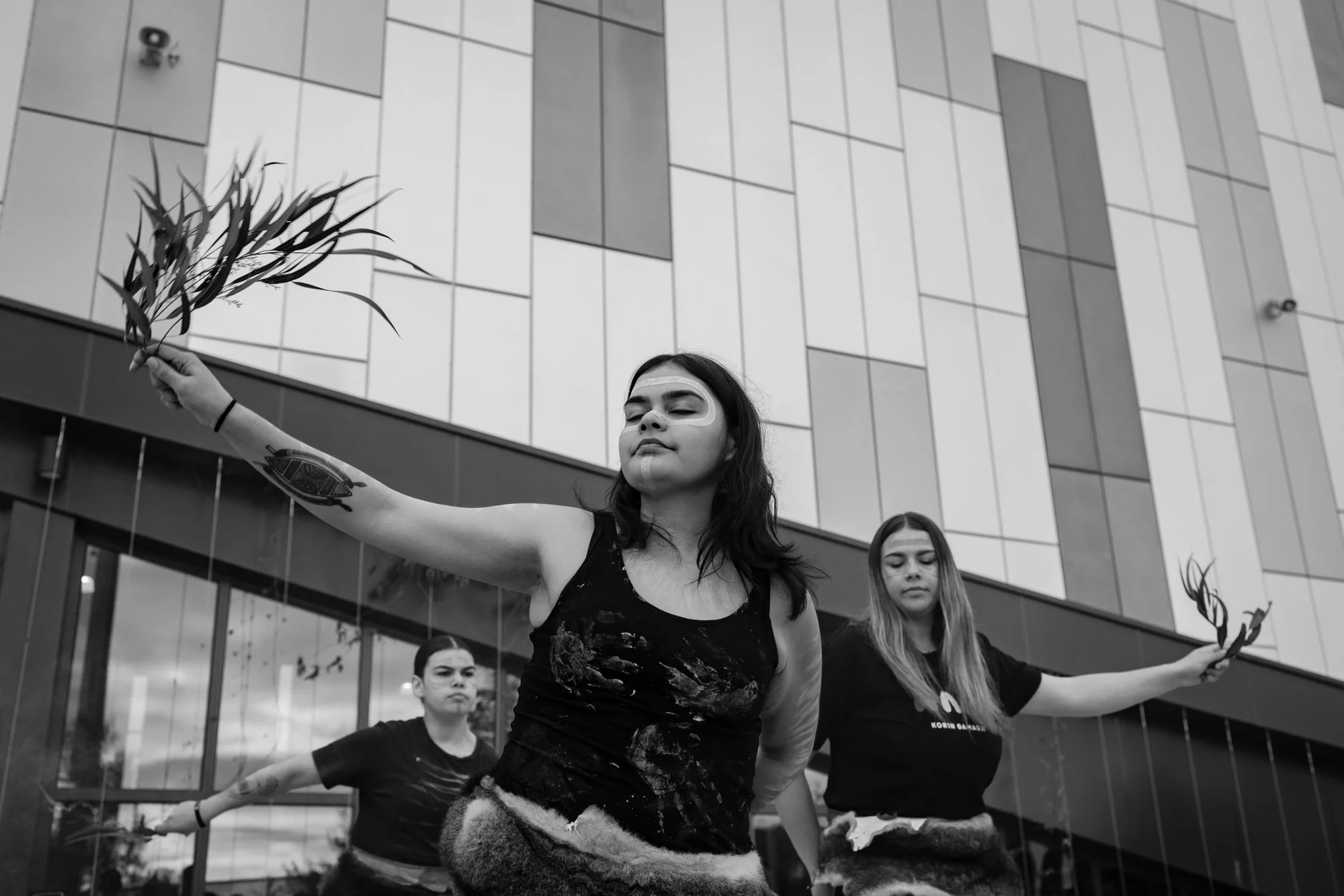 Three indigenous women performing a traditional dance outdoors against a modern building. They have face paint and are holding gum branches in their hands.