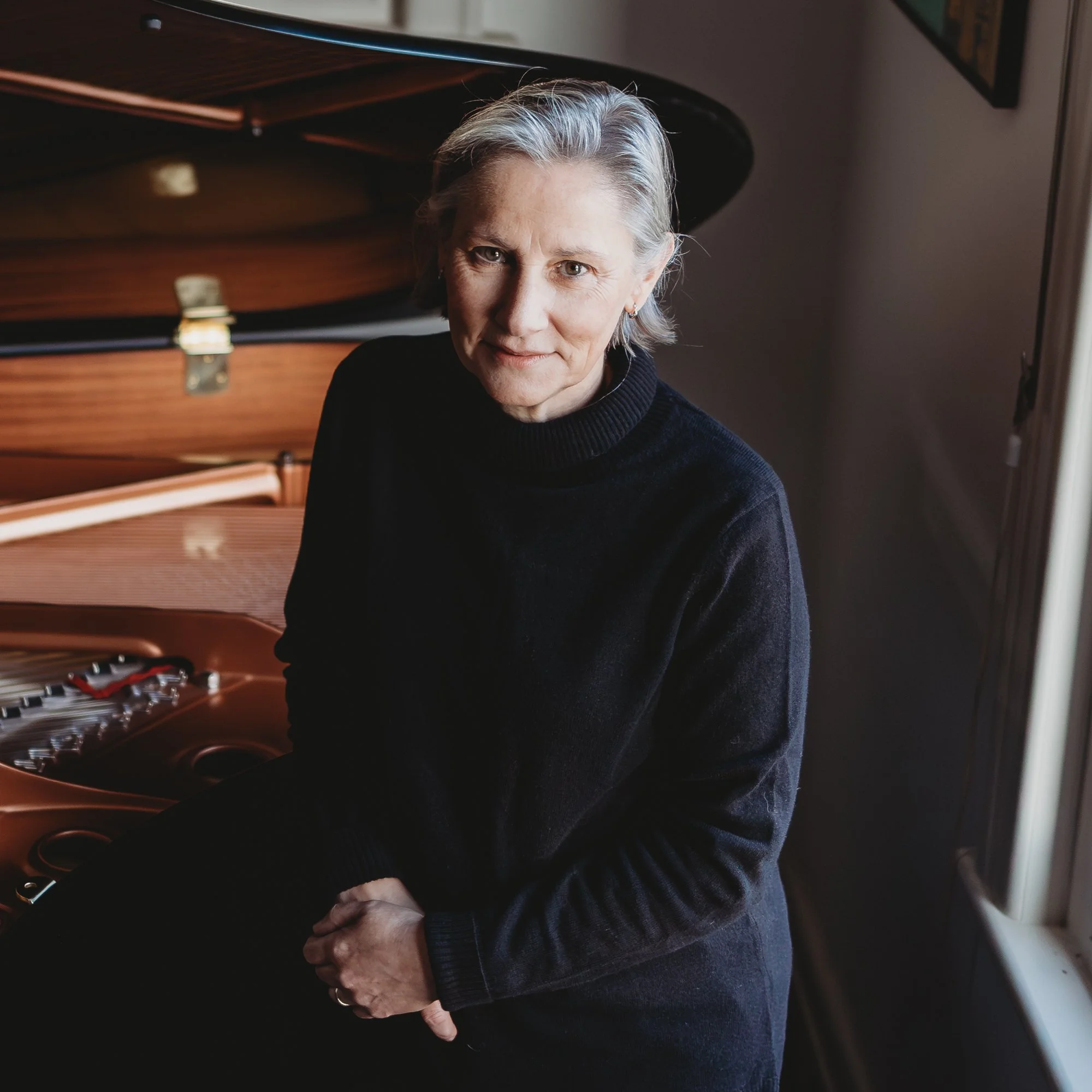 A woman with short gray hair sitting next to a grand piano, wearing a black turtleneck sweater, inside a room with soft lighting.