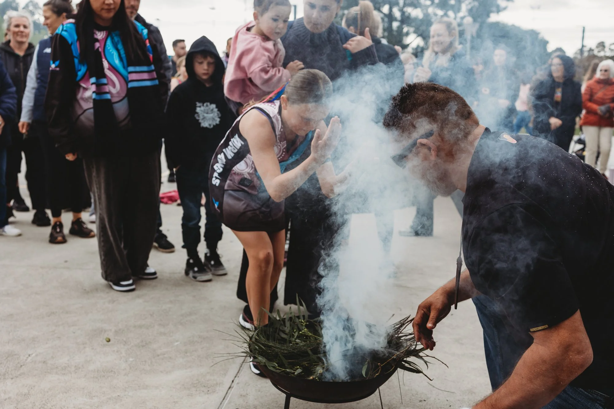 A group of people participate in a traditional smoking ceremony outdoors, with a man tending a fire in a bowl and others observing.