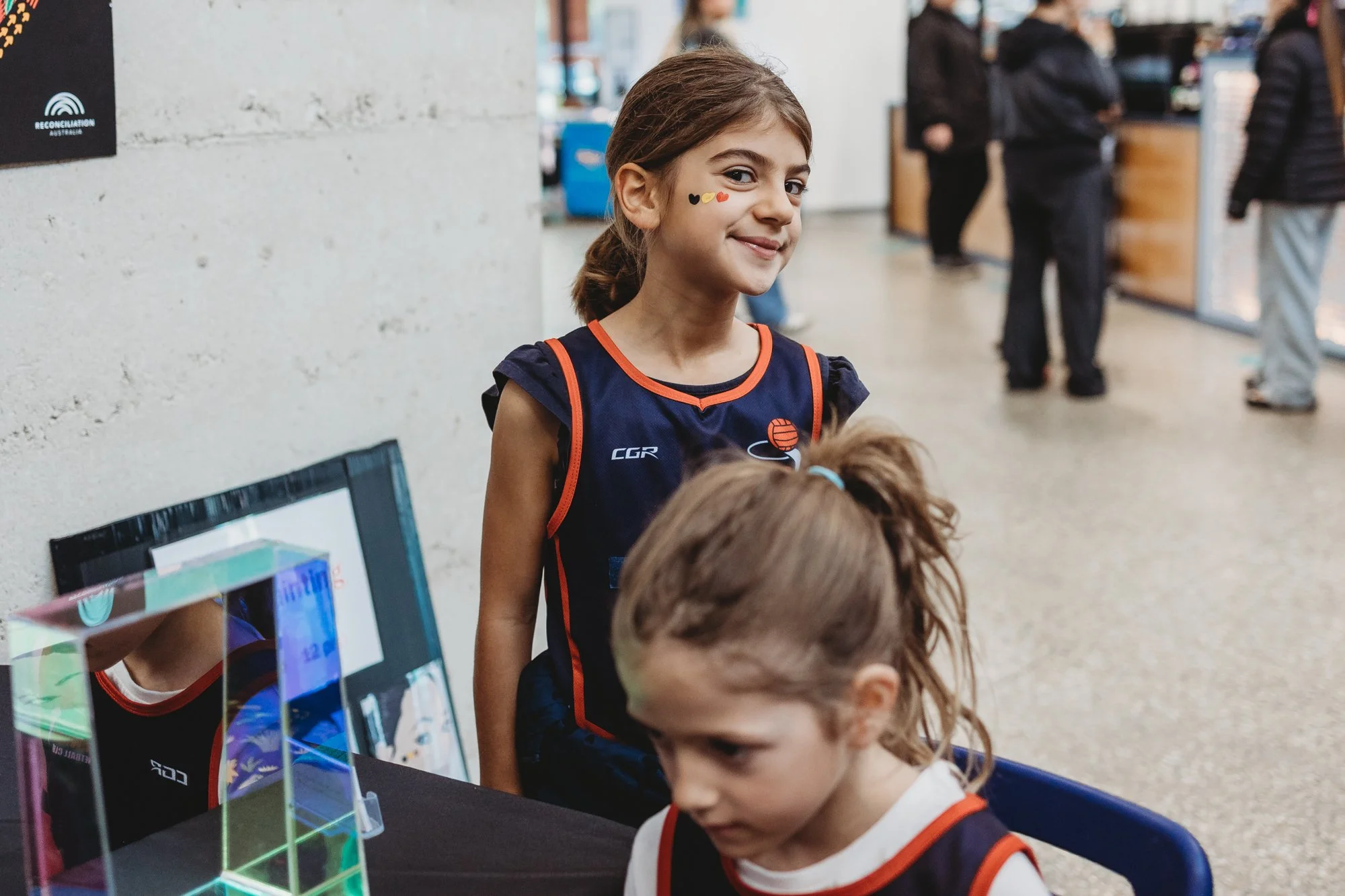 Two young girls wearing netball uniforms, getting indigenous coloured face paint, at a community event.