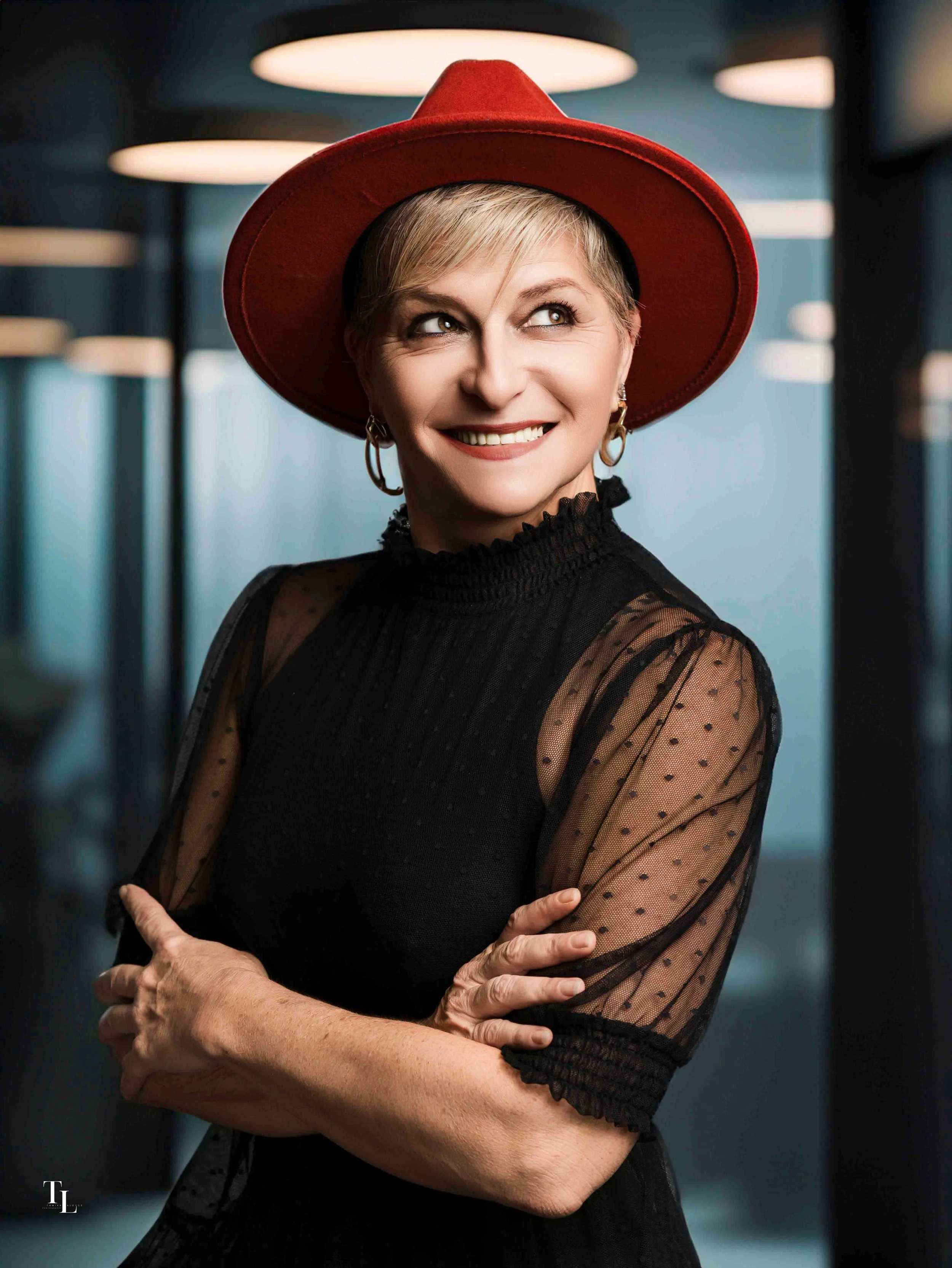 Close-up portrait of a smiling middle-aged woman with short blonde hair, wearing a wide-brimmed red hat and black dress with sheer polka-dot sleeves, standing indoors with blurred background lighting.
