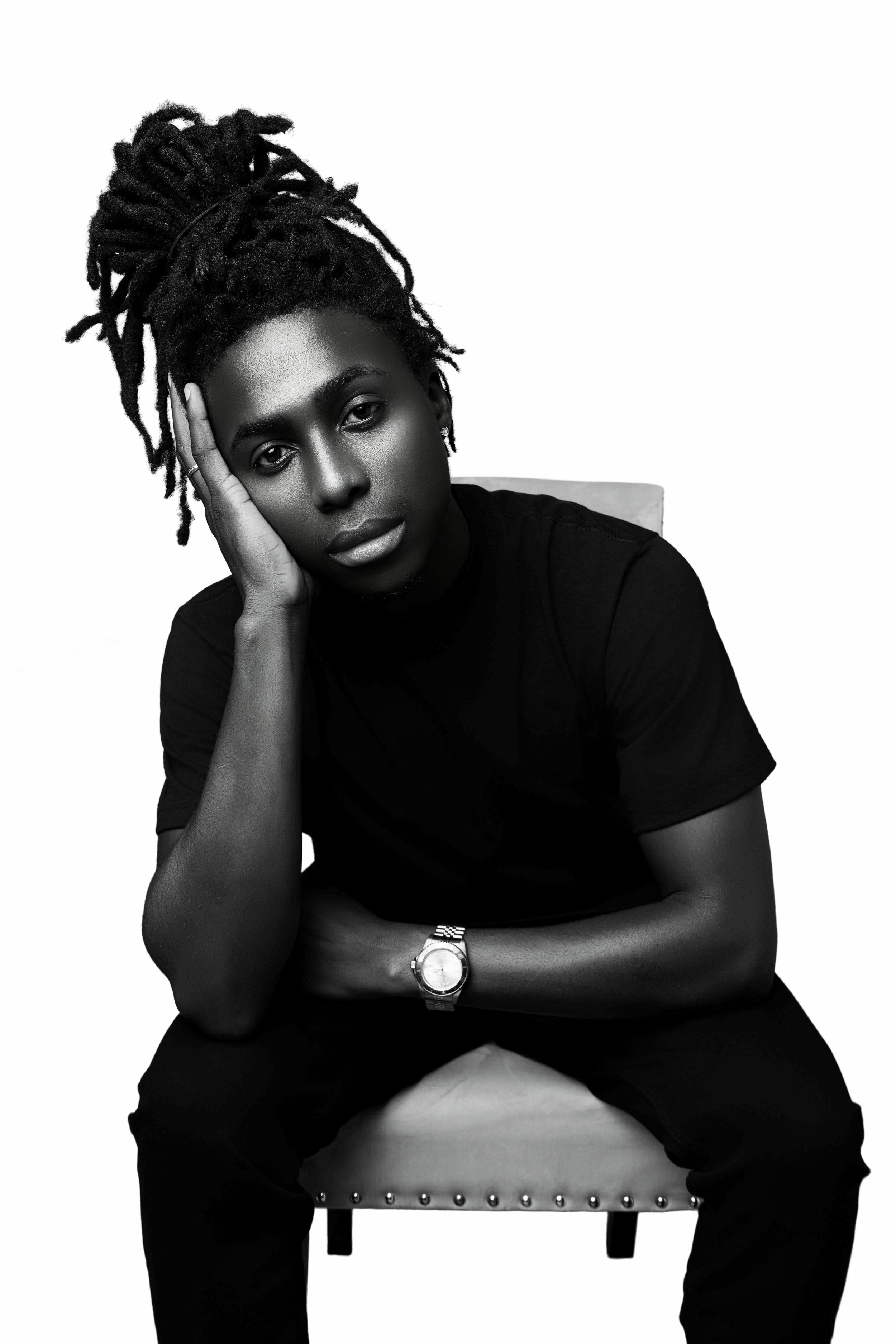 Black and white portrait of a woman with dreadlocks, resting her face on her hand, seated on a chair, wearing a black shirt and a watch.