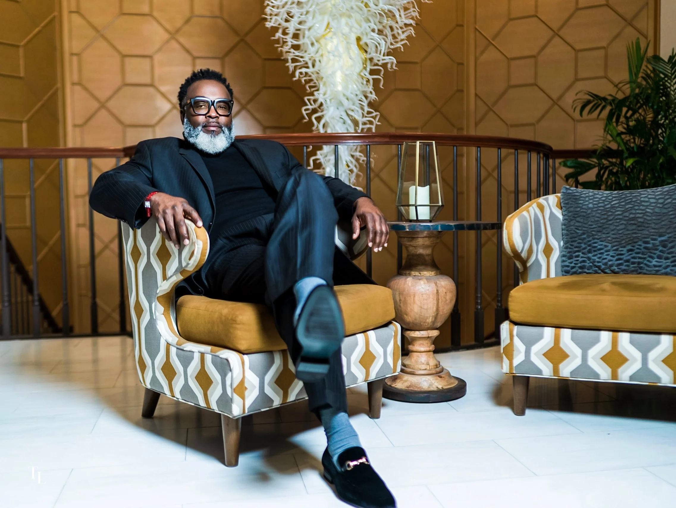 A man with gray beard wearing black glasses and a pinstripe suit sitting on a patterned armchair in a luxurious interior, with a decorative object with white curly elements and a candle behind him.