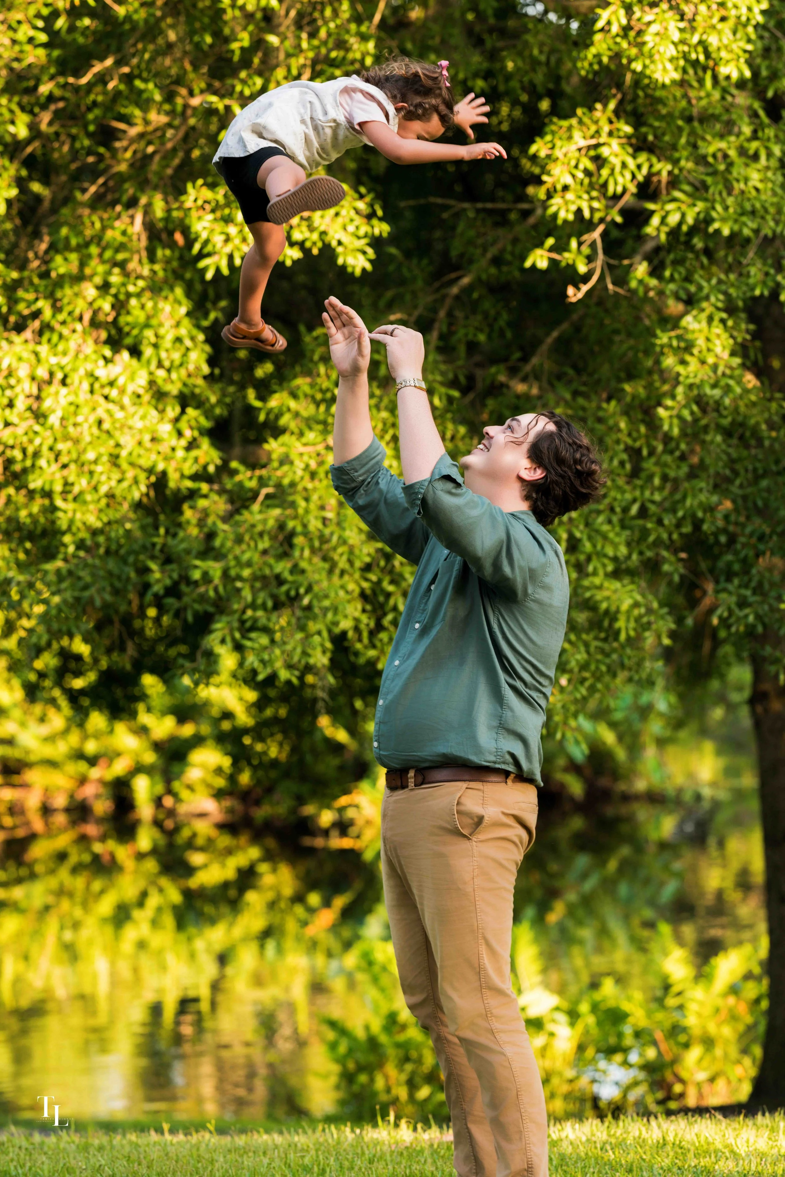 A man with dark, curly hair, wearing a green shirt and khaki pants, is lifting a young girl in the air outdoors in a park or garden with green trees and grass in the background. The girl is wearing a light-colored vest, black shorts, and sandals, and