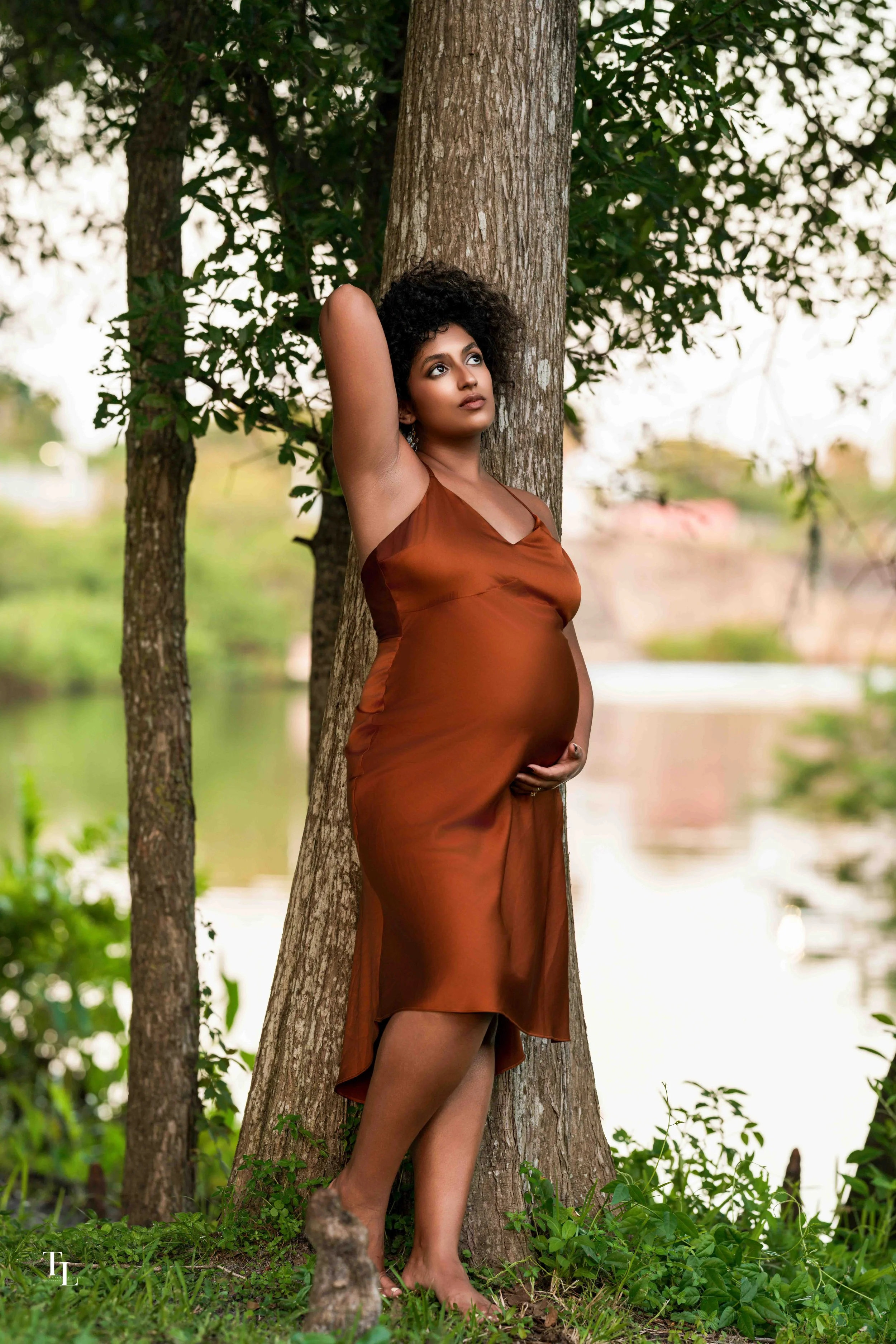 A pregnant woman with curly hair wearing a brown satin dress, standing outdoors by a tree near a body of water, looking thoughtfully into the distance.