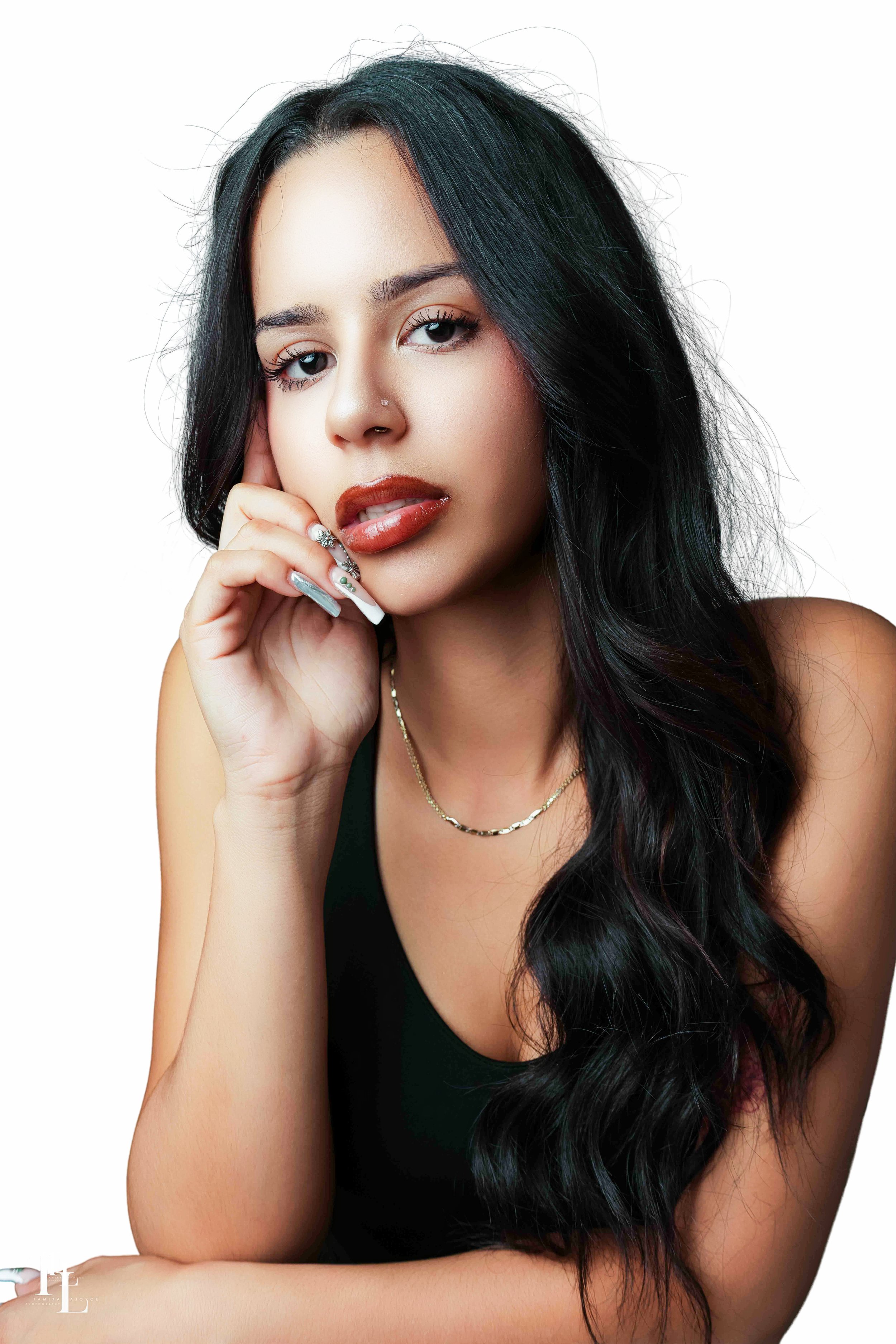 Close-up portrait of a young woman with long, wavy black hair, wearing a black top, with jewelry including rings and necklaces, posing with one hand gently touching her face, against a plain white background.