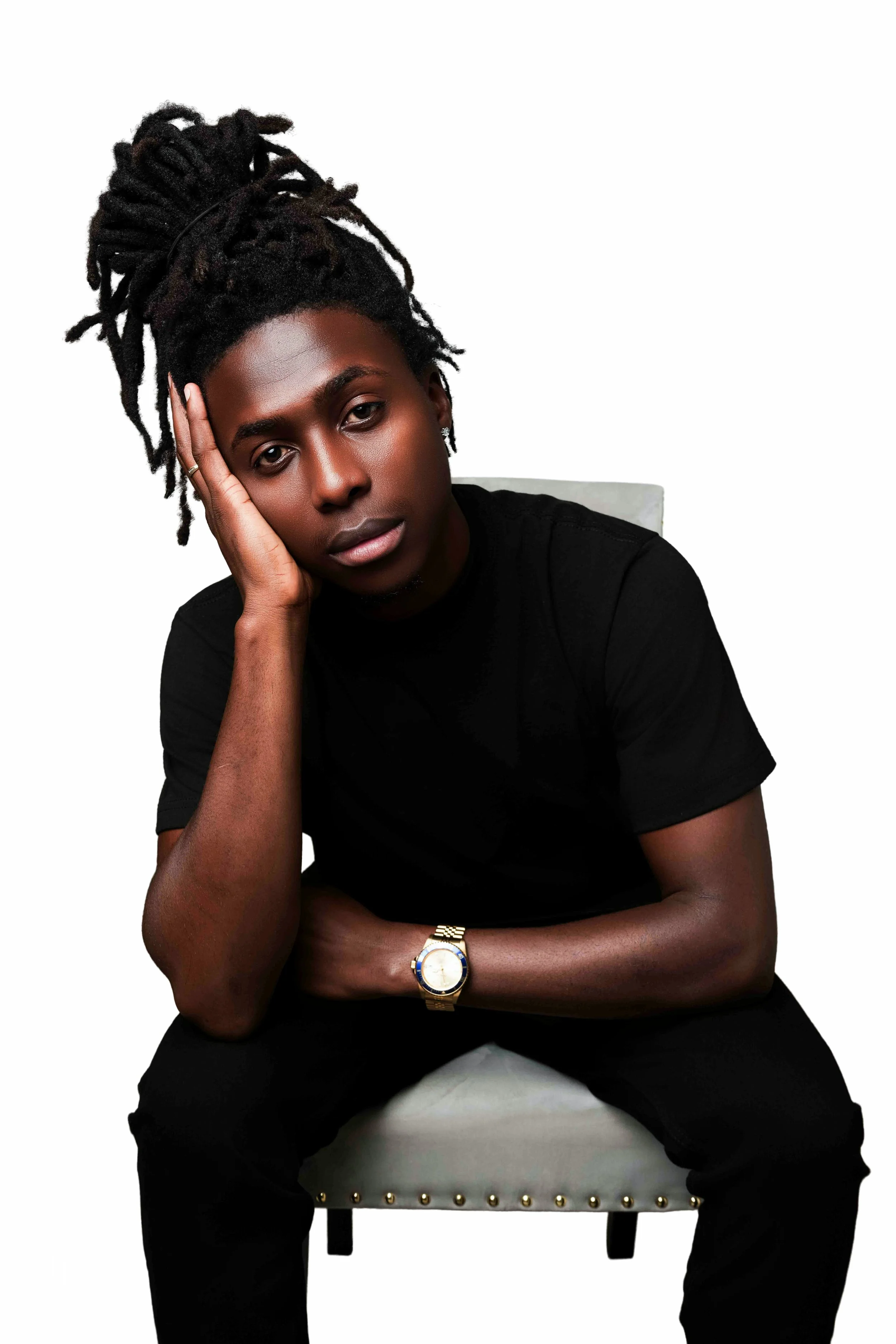A young Black man with dreadlocks sitting on a light-colored chair with brass nailhead details, wearing a black t-shirt, gold watch, and a ring, resting his head on his left hand, looking at the camera against a white background.
