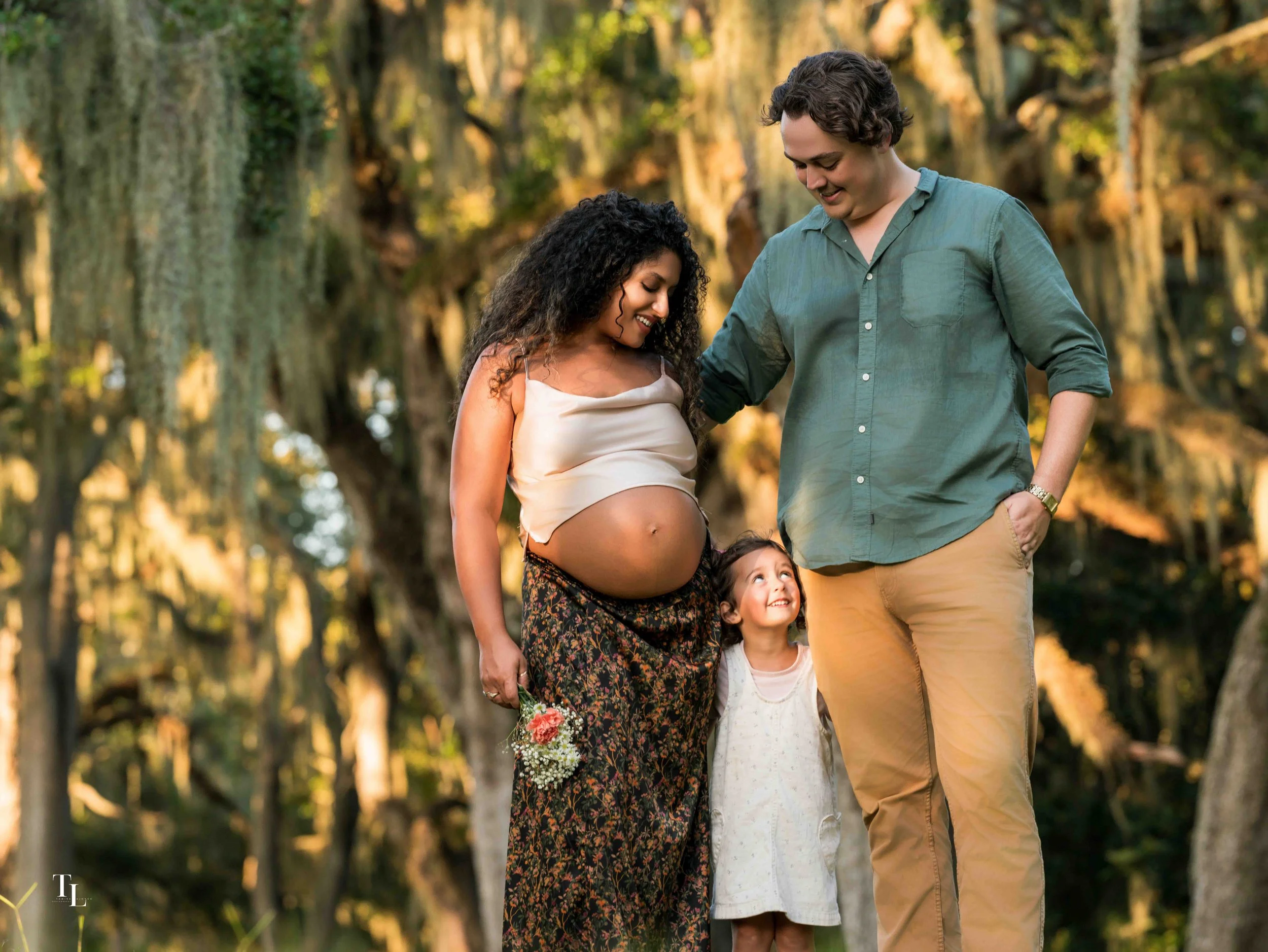 A diverse pregnancy photo of four people, including a pregnant woman, a man, and two young girls, in an outdoor forest setting with trees and sunlight, smiling and interacting.