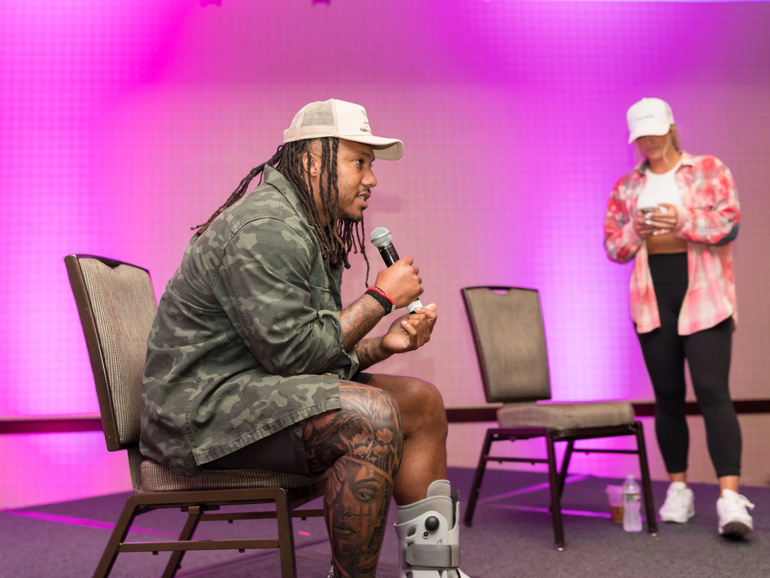 A man with dreadlocks and tattoos sitting on a chair, speaking into a microphone at an event, with a woman standing in the background looking at her phone against a pink backdrop.