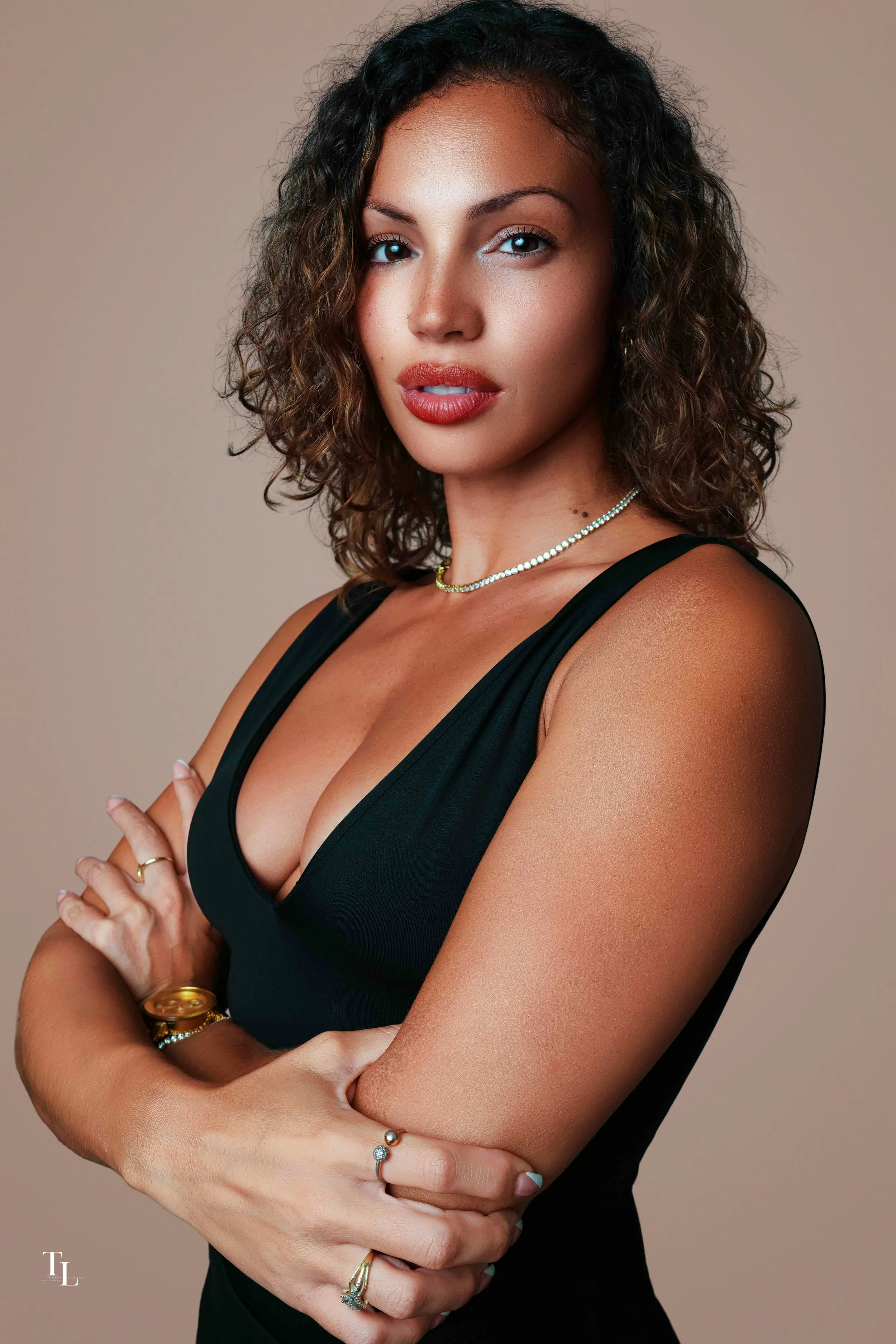 A woman with curly brown hair and blue eyes wearing a black sleeveless top, multiple rings, bracelets, a necklace, and light makeup, looking confidently at the camera with arms crossed.