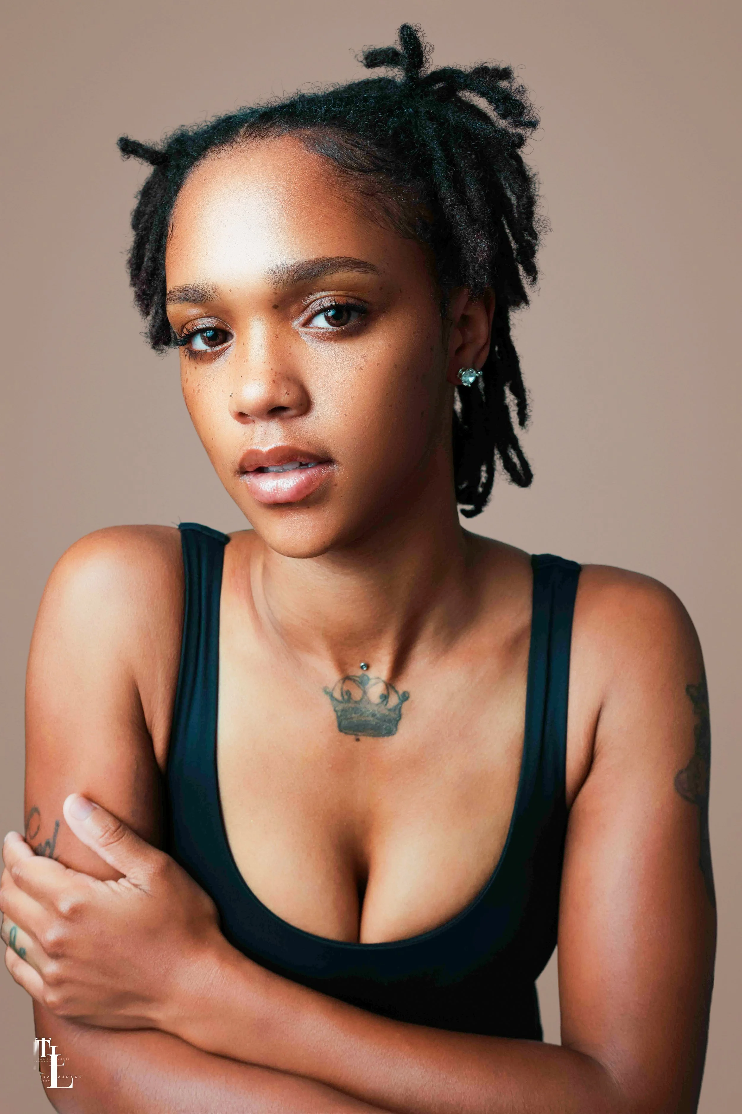 Portrait of a woman with short, curly black hair, wearing a black tank top, adorned with earrings, and a tattoo of a crown on her chest. She is looking at the camera with a neutral expression against a plain background.