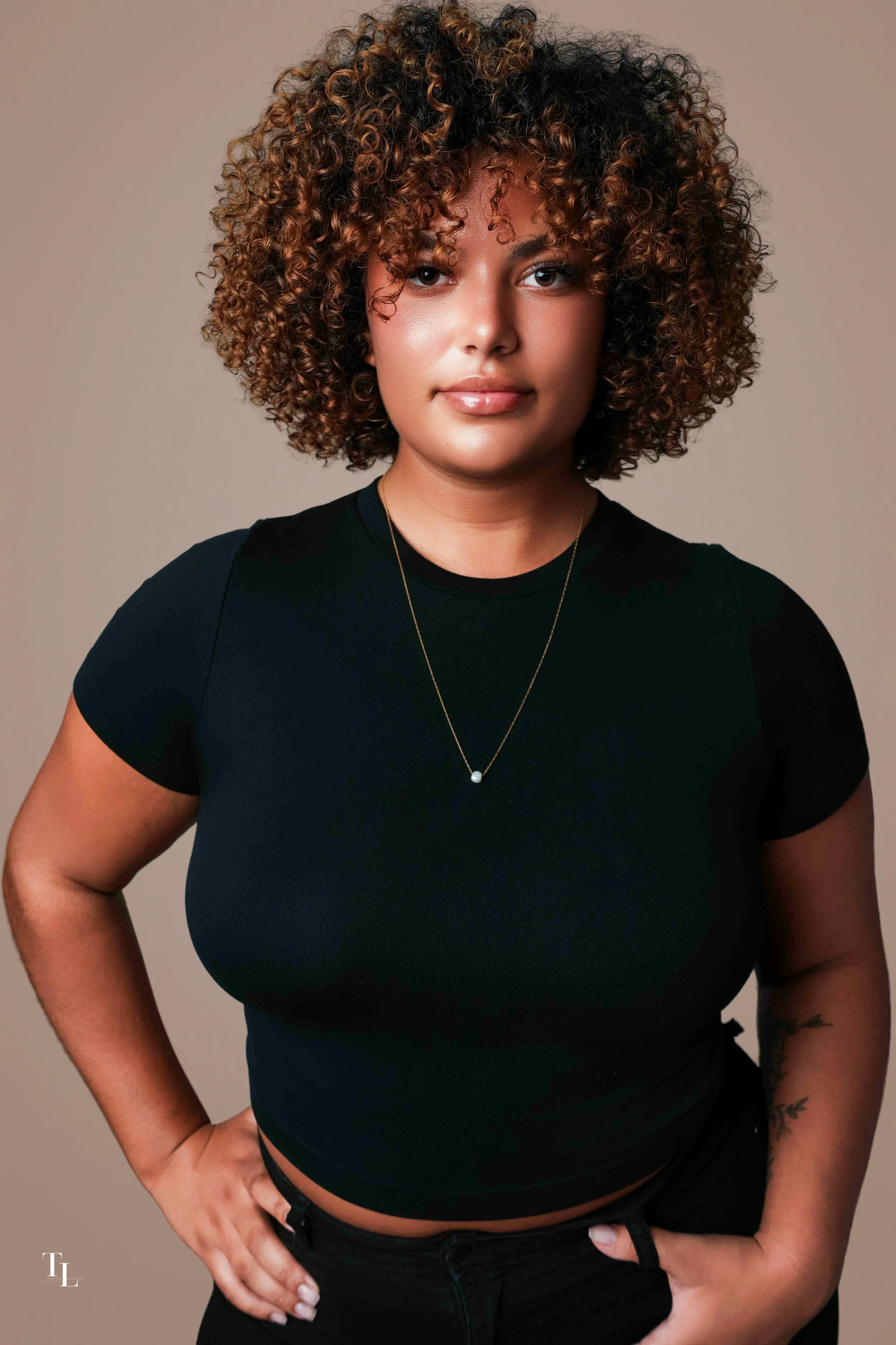A woman with curly, shoulder-length hair wearing a black t-shirt with a round neckline, a necklace with a pearl pendant, and black pants. She is standing against a plain neutral background, with her right hand on her hip and her left hand resting on 