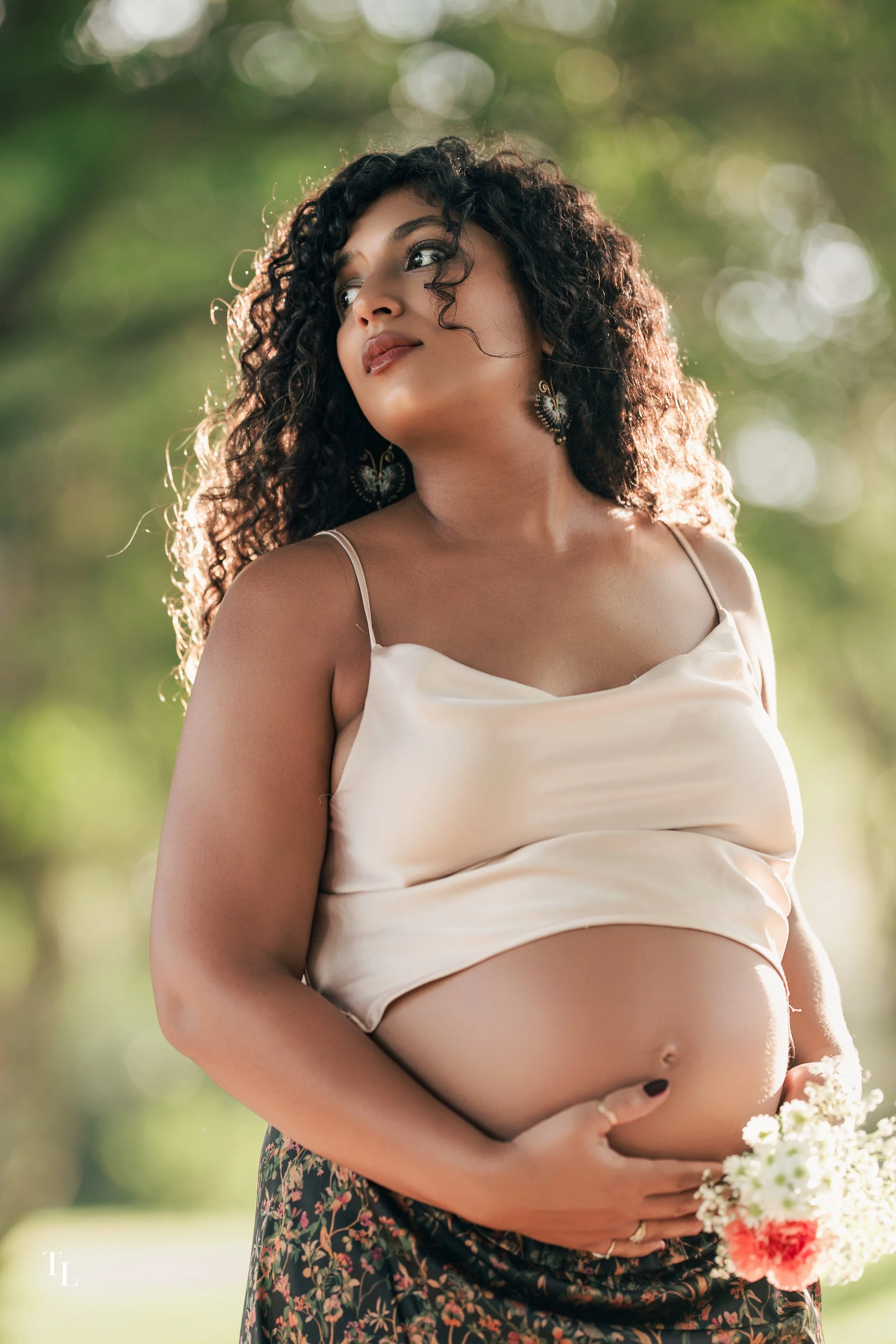A pregnant woman with curly dark hair wearing a satin-like cream-colored top and floral skirt, holding a bouquet of flowers outdoors against a blurred green background.
