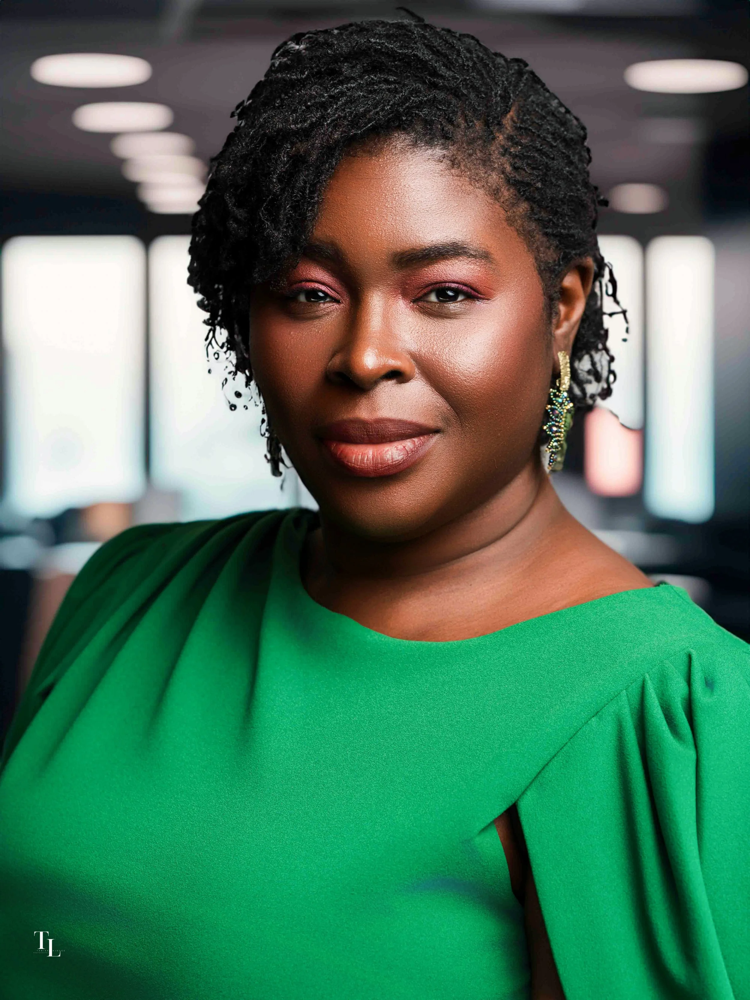 Close-up of a woman with dark skin and short, twisted black hair, wearing a bright green dress and colorful earrings, in a modern indoor setting.