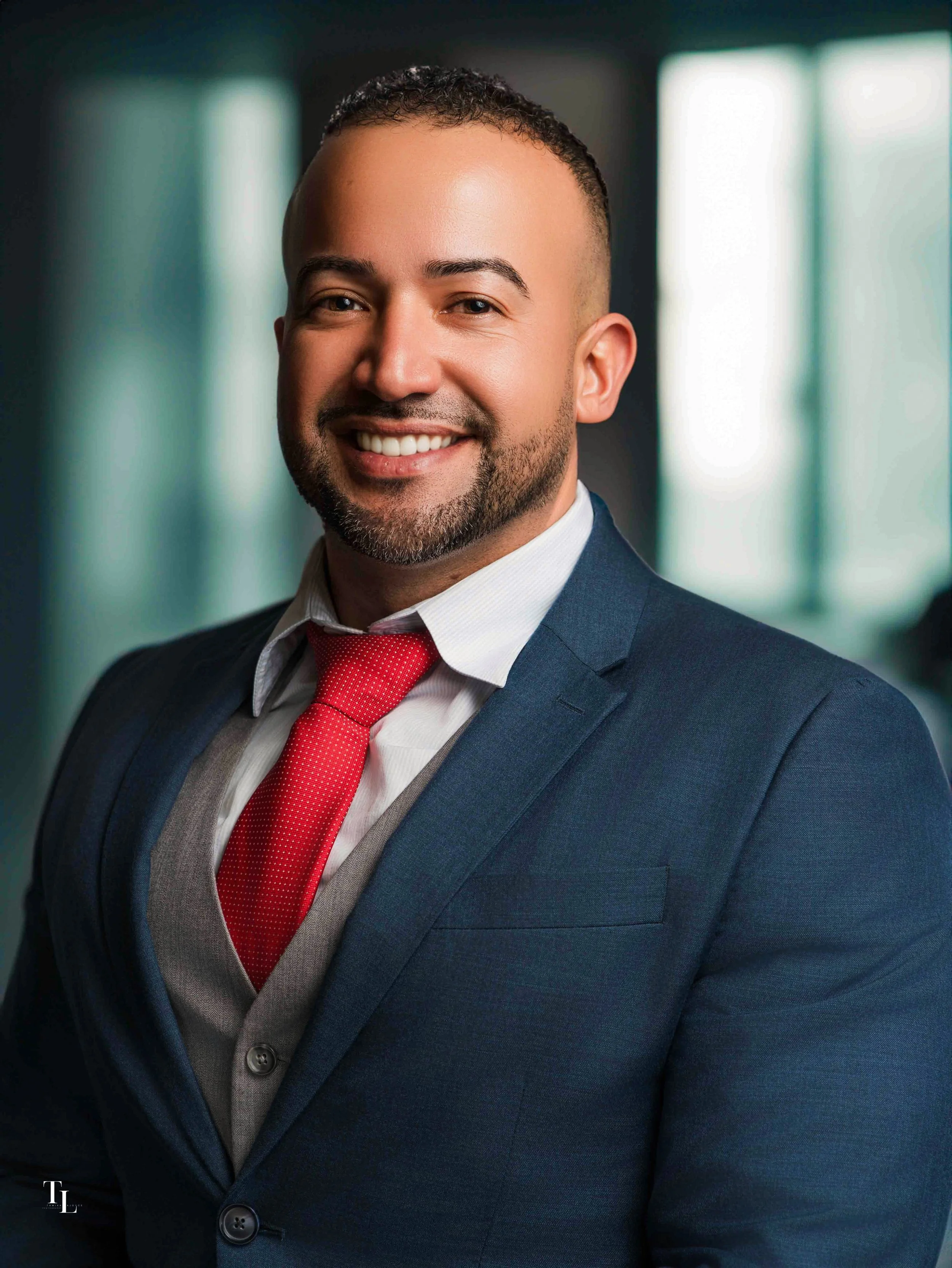 Portrait of a smiling man with a neatly groomed beard and short curly hair, wearing a navy blue suit, a white shirt, and a red tie, standing in a professional setting with blurred windows in the background.