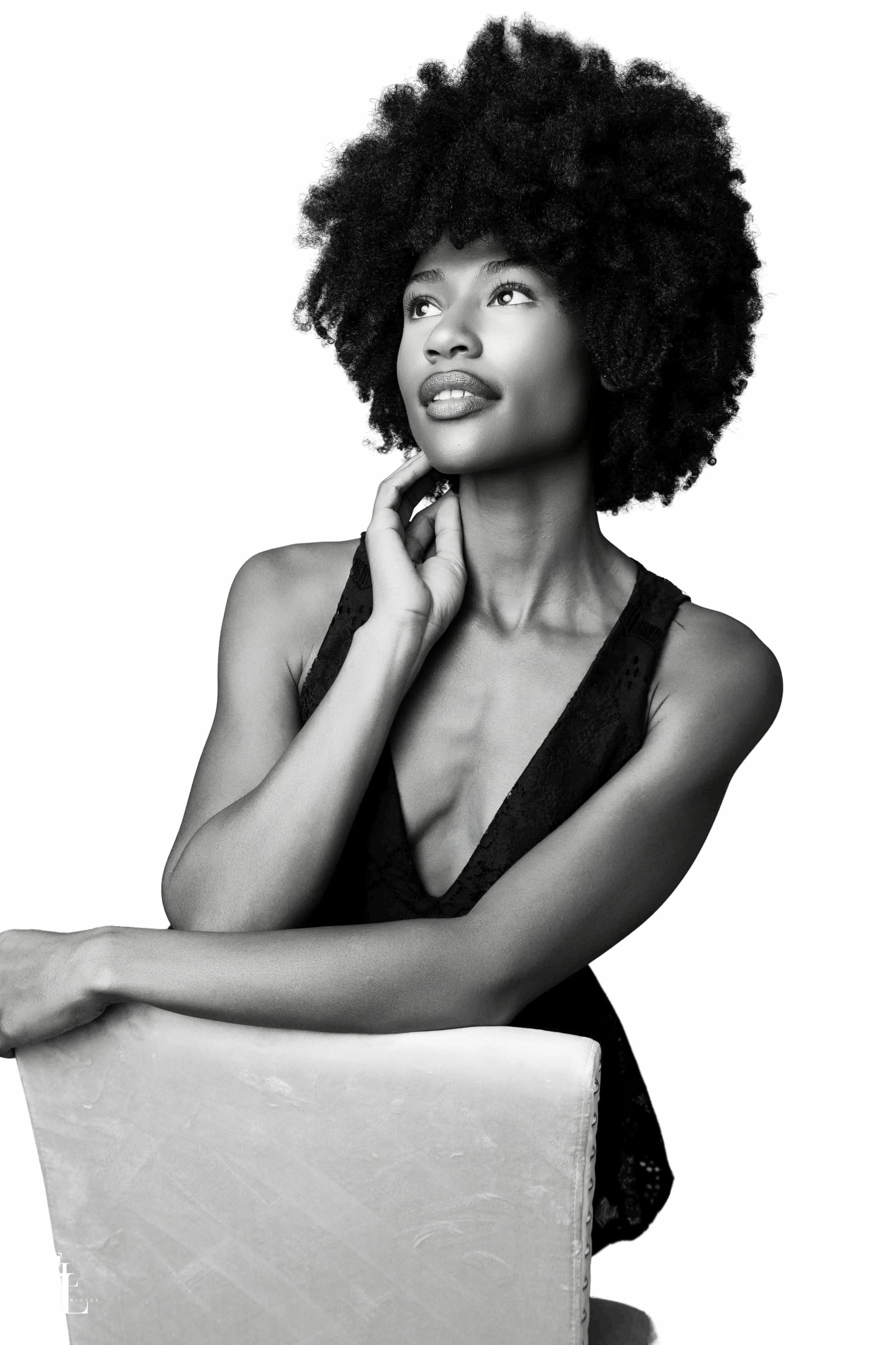 Black and white photograph of a woman with natural curly hair, looking upwards, resting her chin on her hand, wearing a sleeveless top.