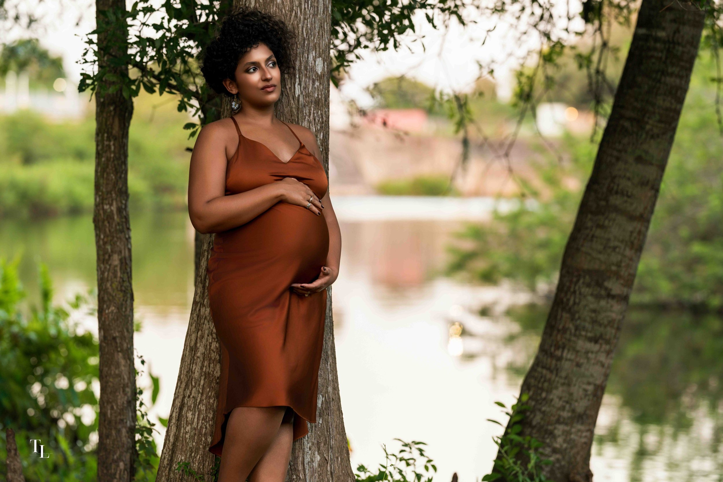 A pregnant woman standing by a tree near a body of water in a lush green setting, wearing a brown dress and jewelry, looking contemplative.