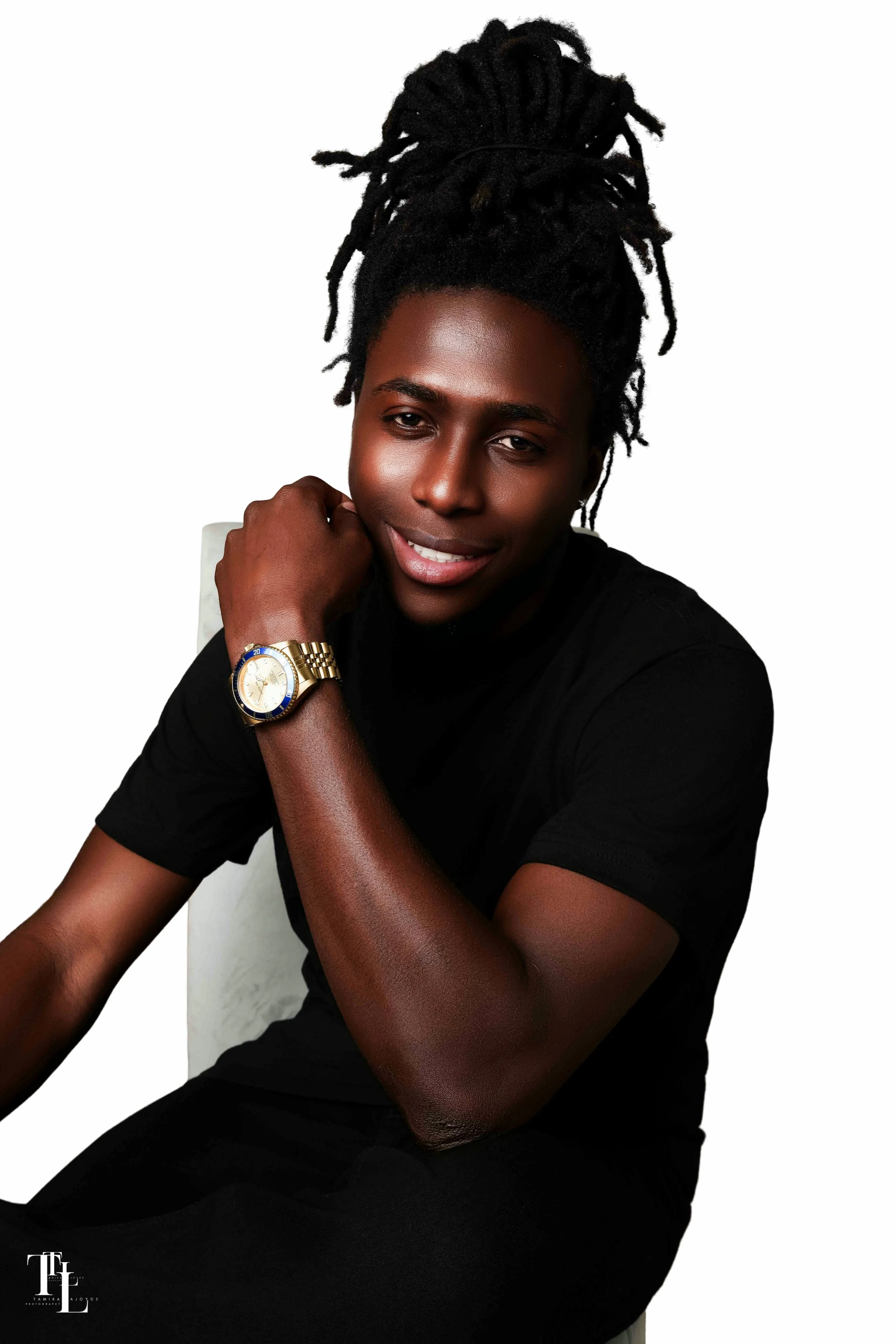 A young woman with dark skin and dreadlocks styled in a high bun, smiling slightly, wearing a black top, a gold wristwatch, and a ring, sitting against a white background.