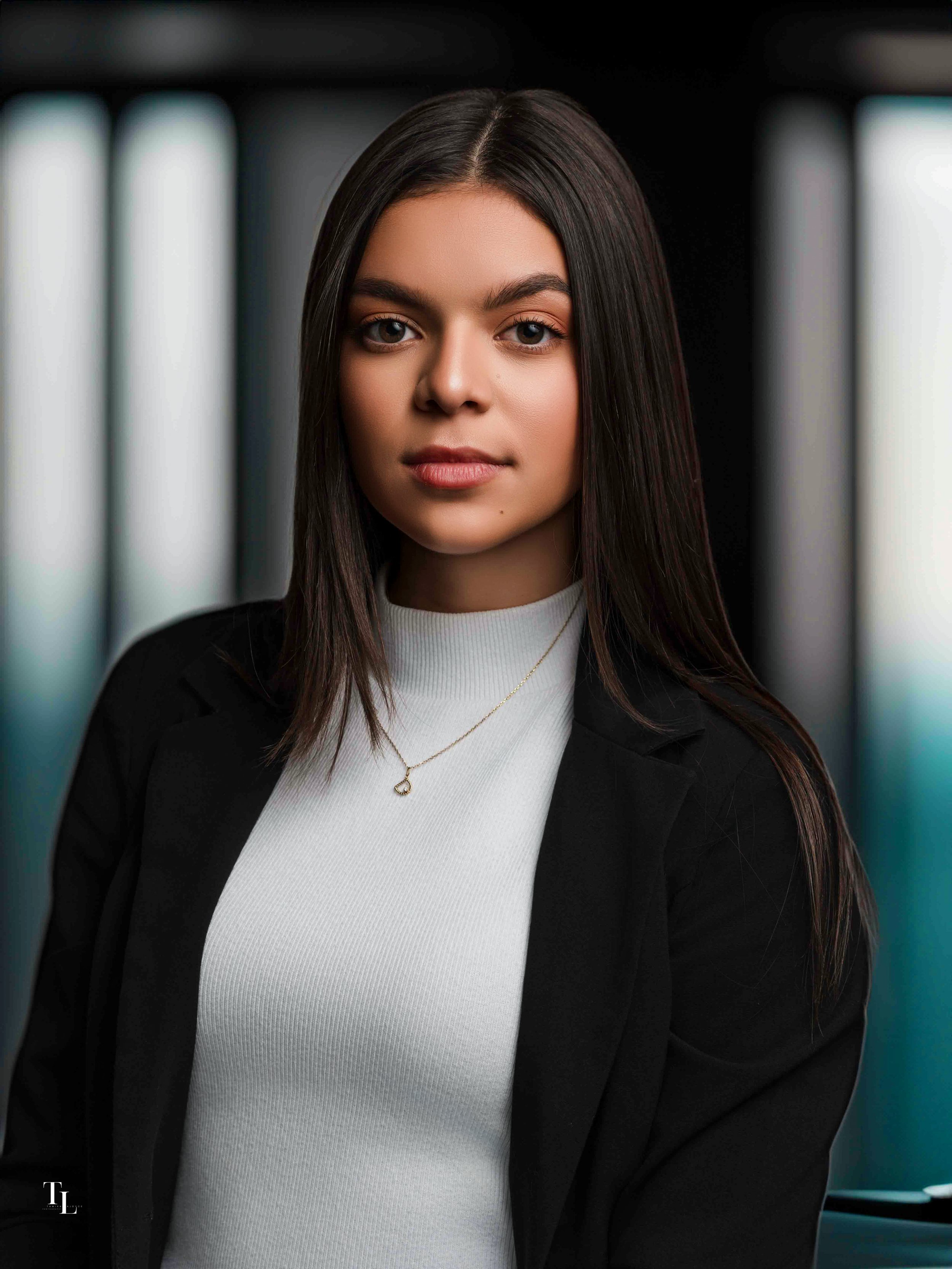 A young woman with long dark hair wearing a white top, black blazer, and a gold heart pendant necklace, standing in front of blurred vertical lights, looking directly at the camera.