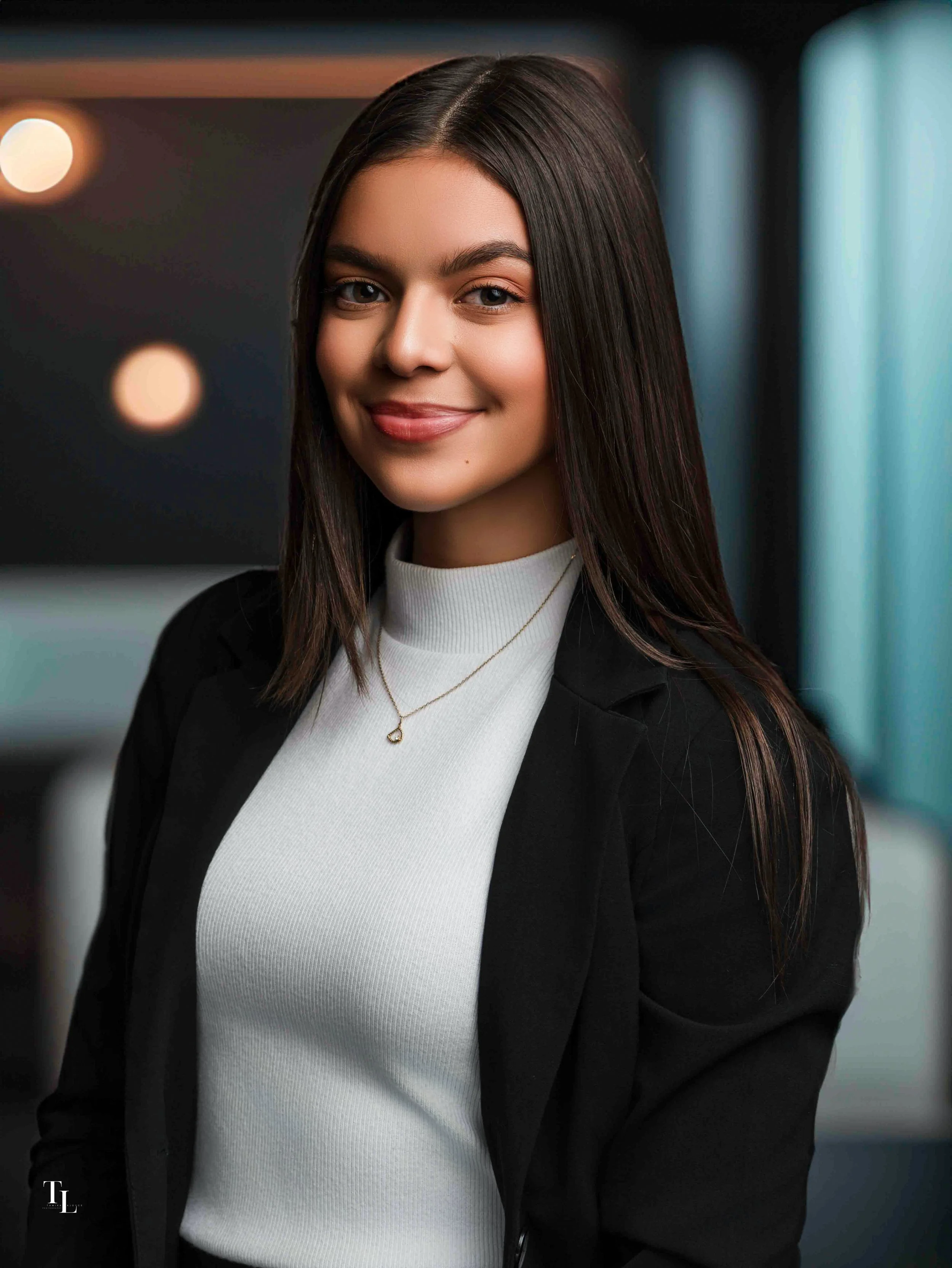 Close-up of a young woman with long dark hair, smiling, wearing a white top and a black jacket, with a blurred modern interior background.