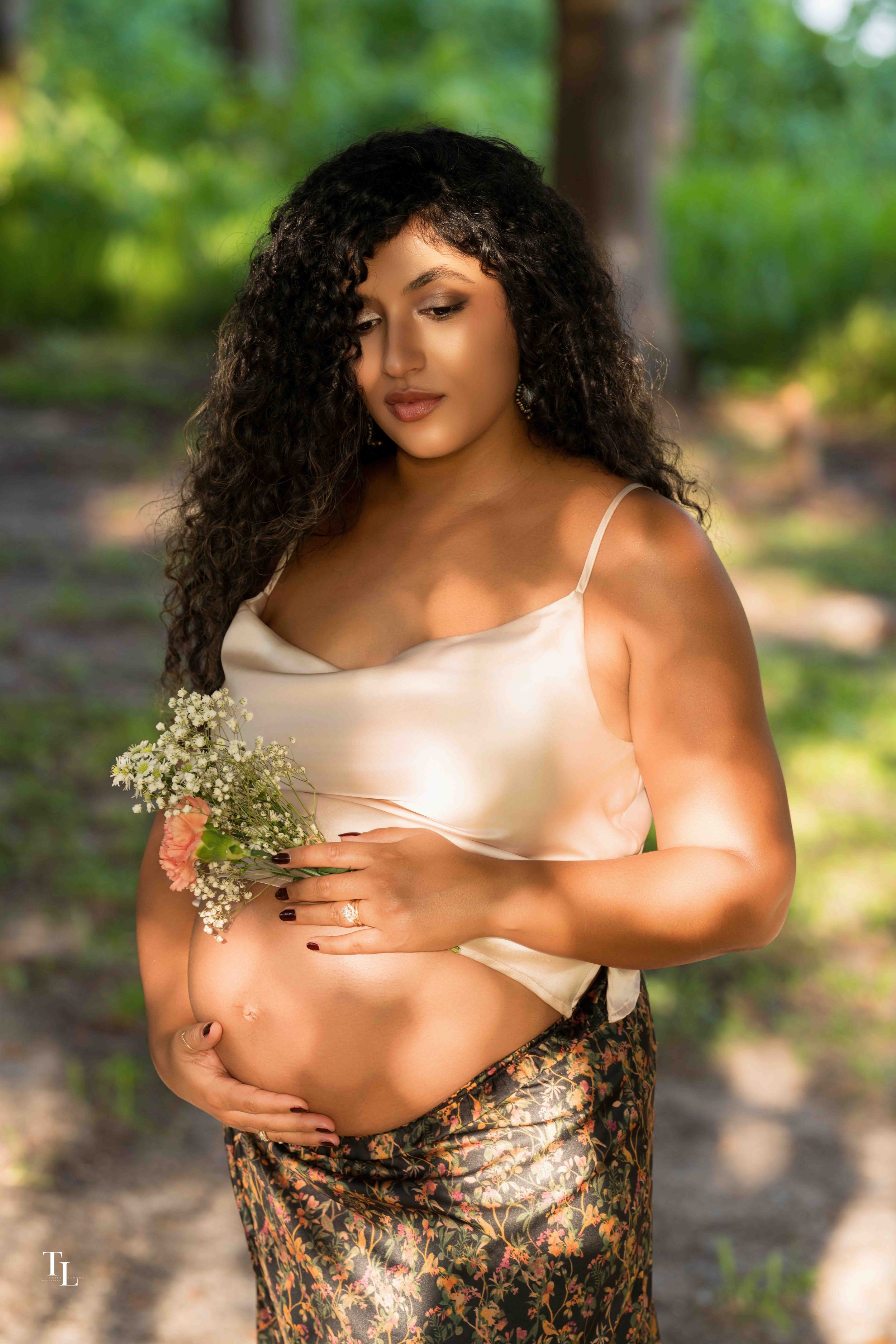 Pregnant woman with curly hair in a white satin top and floral skirt holding a small bouquet of flowers in a forest setting.