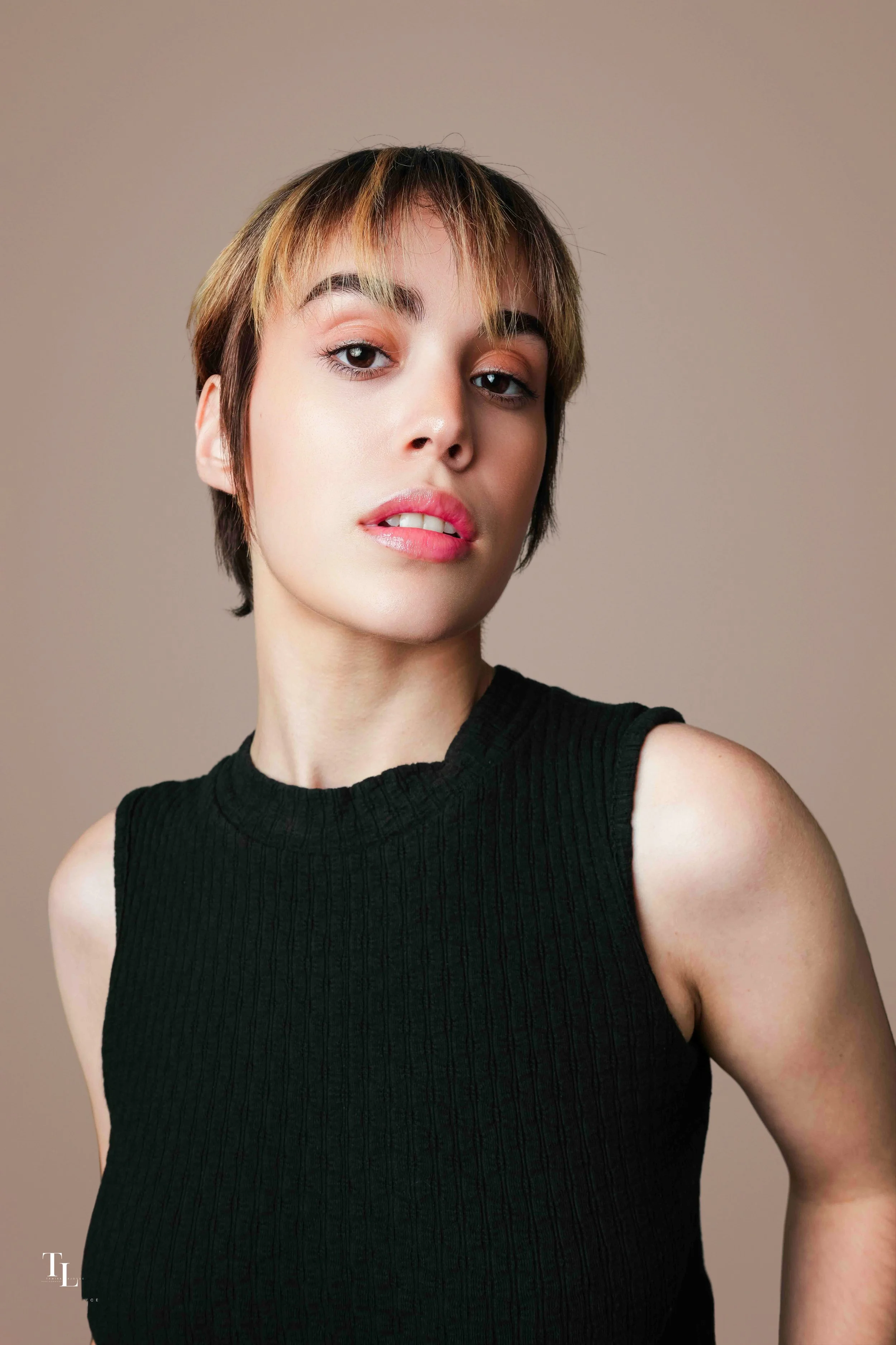 Portrait of a young woman with short, layered haircut and light makeup, wearing a sleeveless black top, against a neutral background.