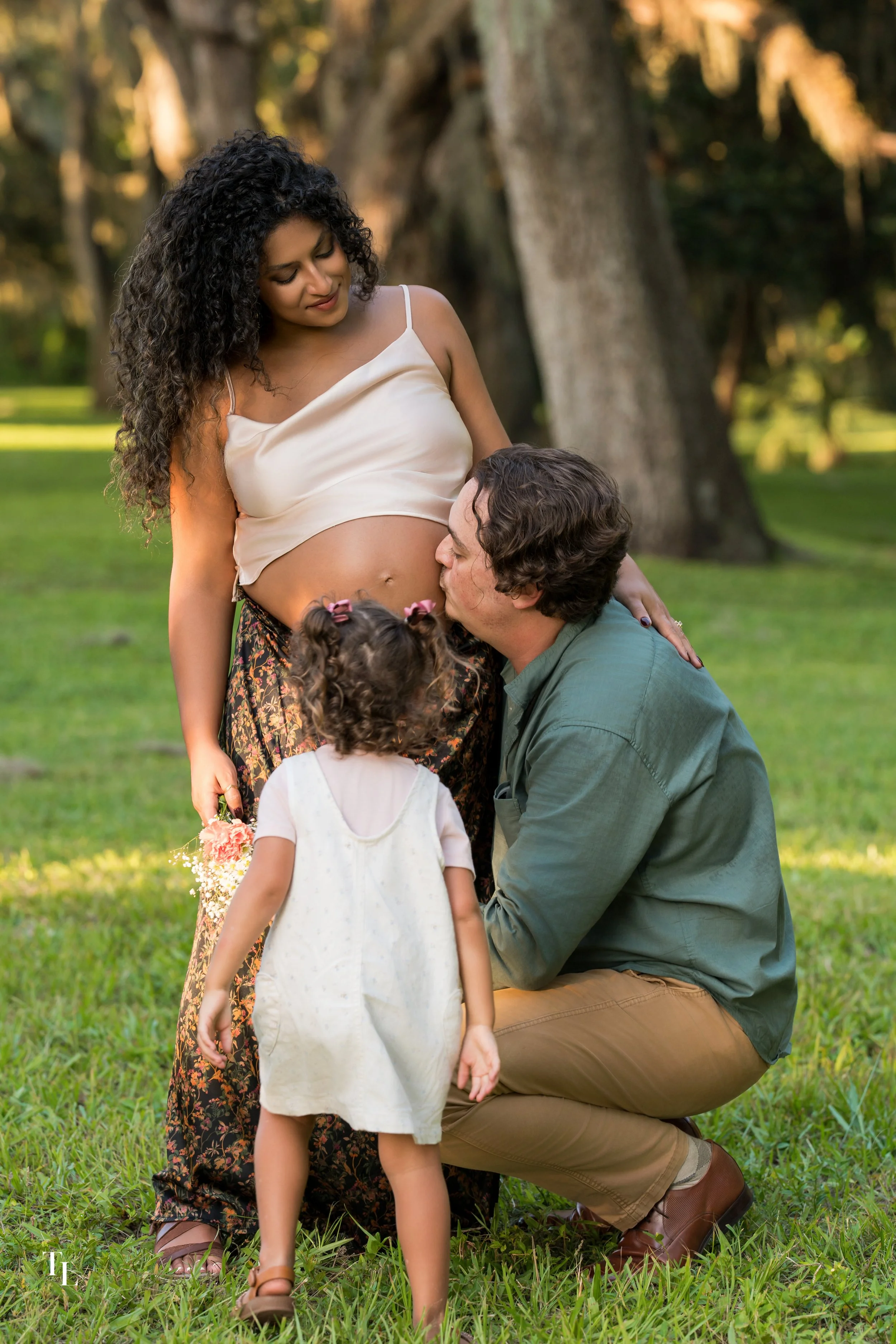 A pregnant woman in a white spaghetti strap top holding a bouquet of flowers, standing outdoors in a park with large trees in the background. A man kneeling and kissing her baby bump, two young girls with curly hair facing away, one in a cream dress 