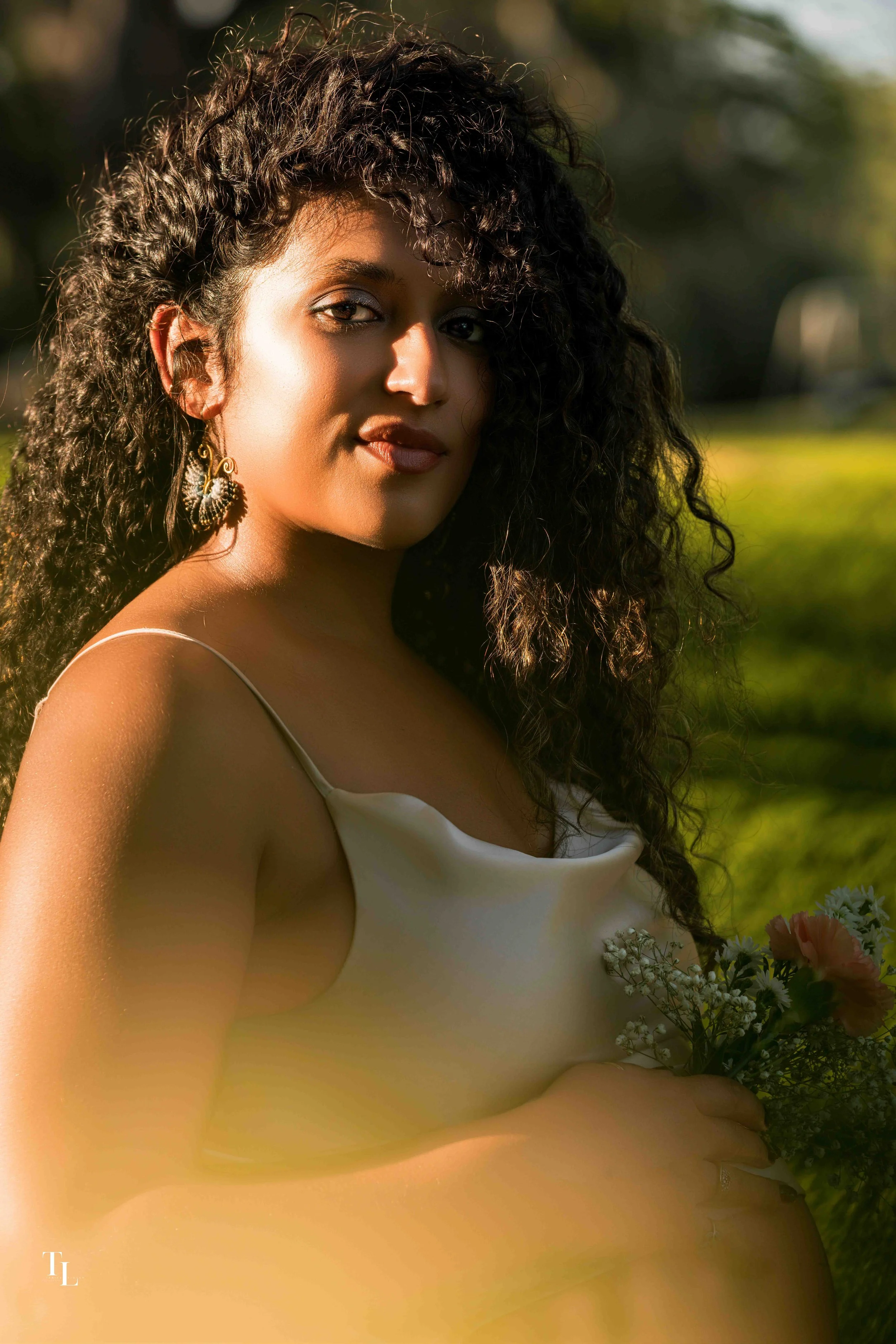 A woman with curly hair and earrings, holding a bouquet of flowers, outdoors with sunlight highlighting her face and shoulder.