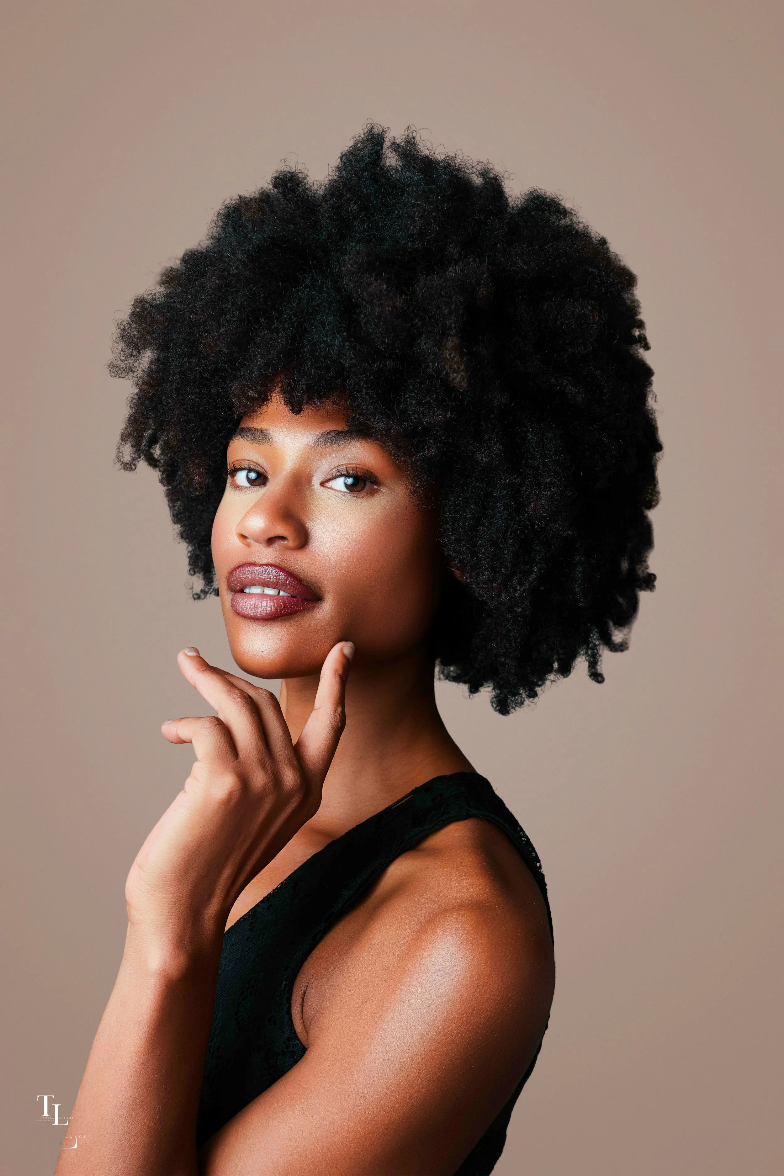 Portrait of a woman with natural curly afro hairstyle, posing with her hand gently touching her chin, wearing a black sleeveless top against a neutral background.