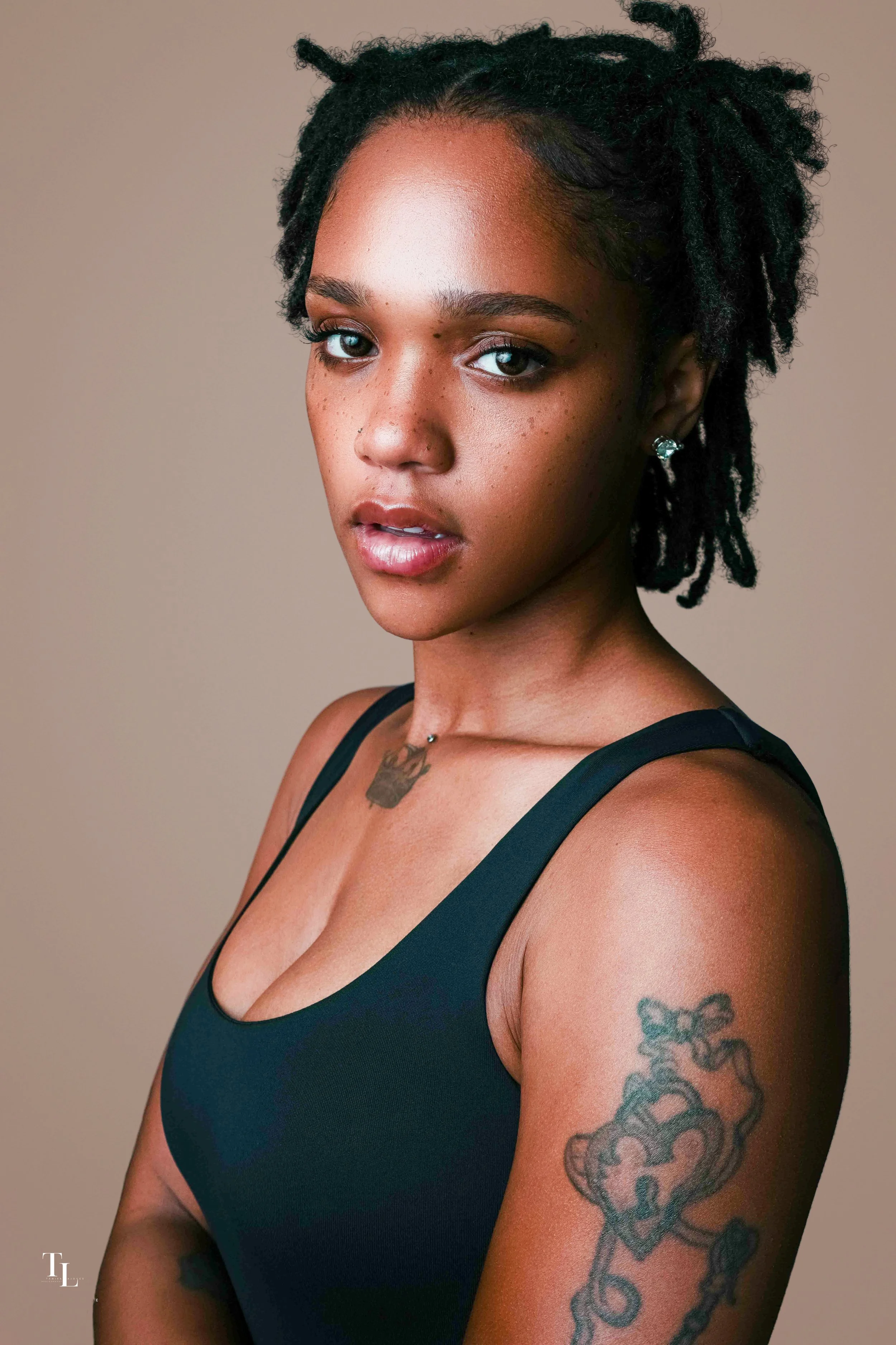 Close-up portrait of a young woman with medium brown skin, dark curly hair styled in twists, blue eyes, and tattoos on her right arm and collarbone, wearing a black tank top, looking directly at the camera against a neutral background.