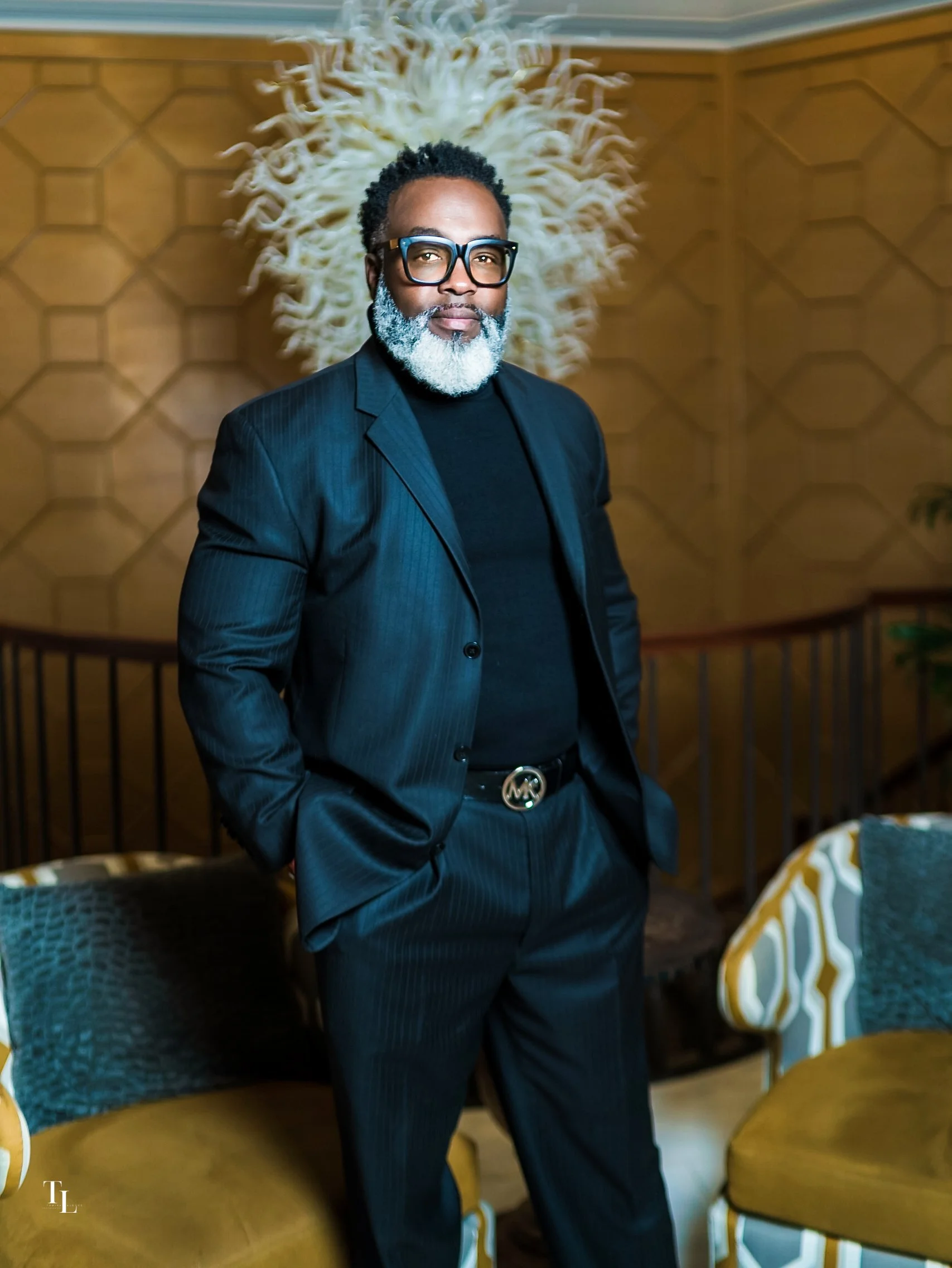 A man with glasses and a gray beard wearing a dark pinstripe suit and black turtleneck, standing in a restaurant or banquet space with honeycomb patterned wooden walls, with tables and chairs around, and a large wooden sculpture of a sea anemone or c