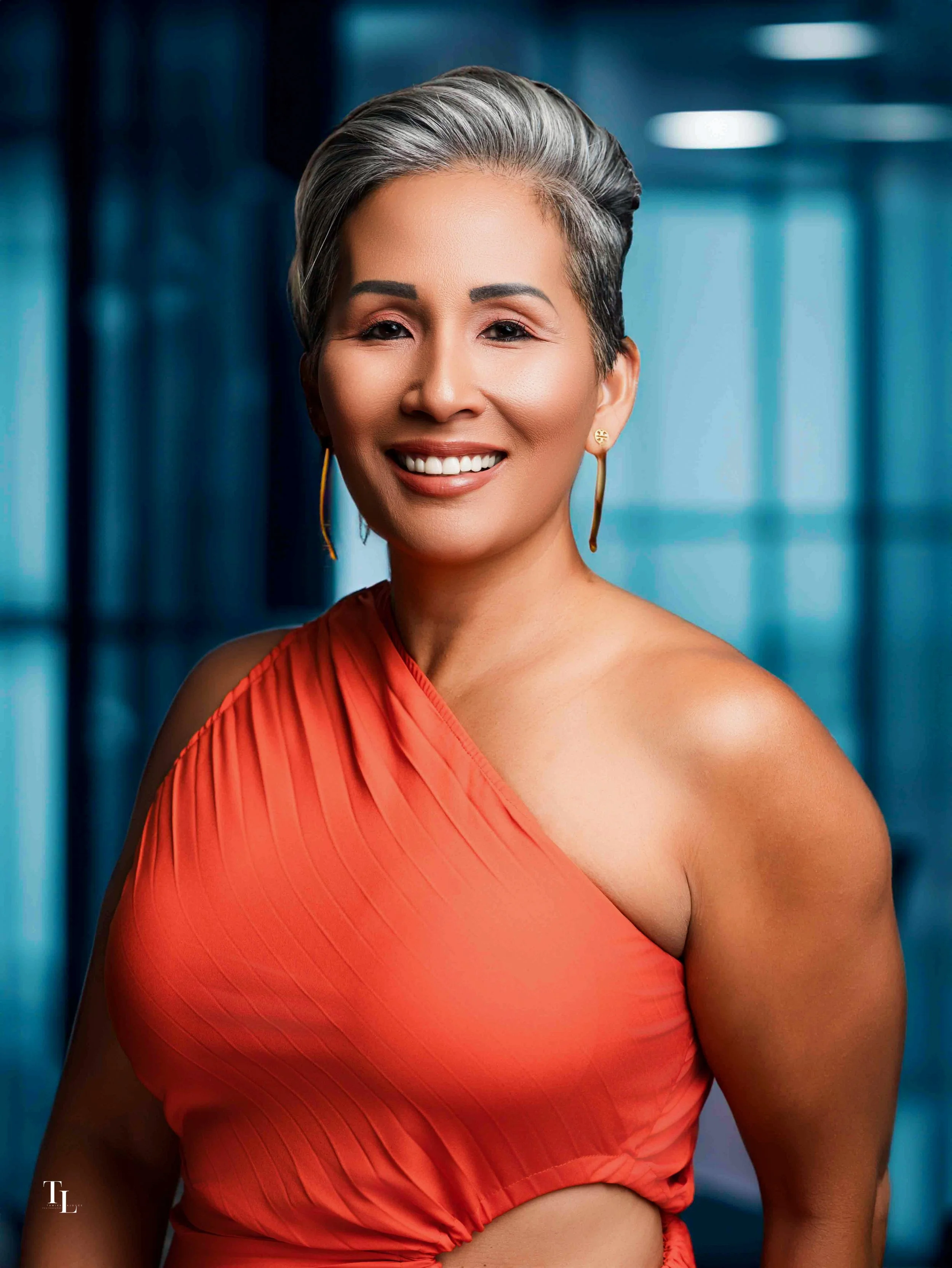 Portrait of a woman with short gray hair wearing a one-shoulder red dress and gold earrings, standing in front of a blurred blue background.