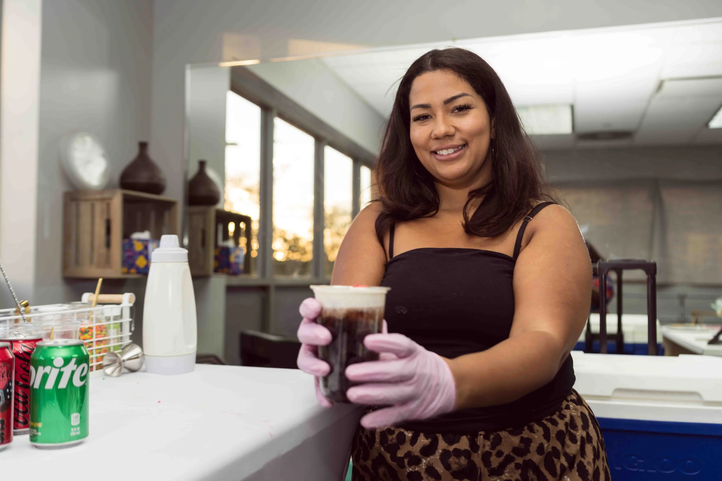 A woman smiling while holding a cup of soda in a classroom or workshop setting with shelves and supplies in the background.
