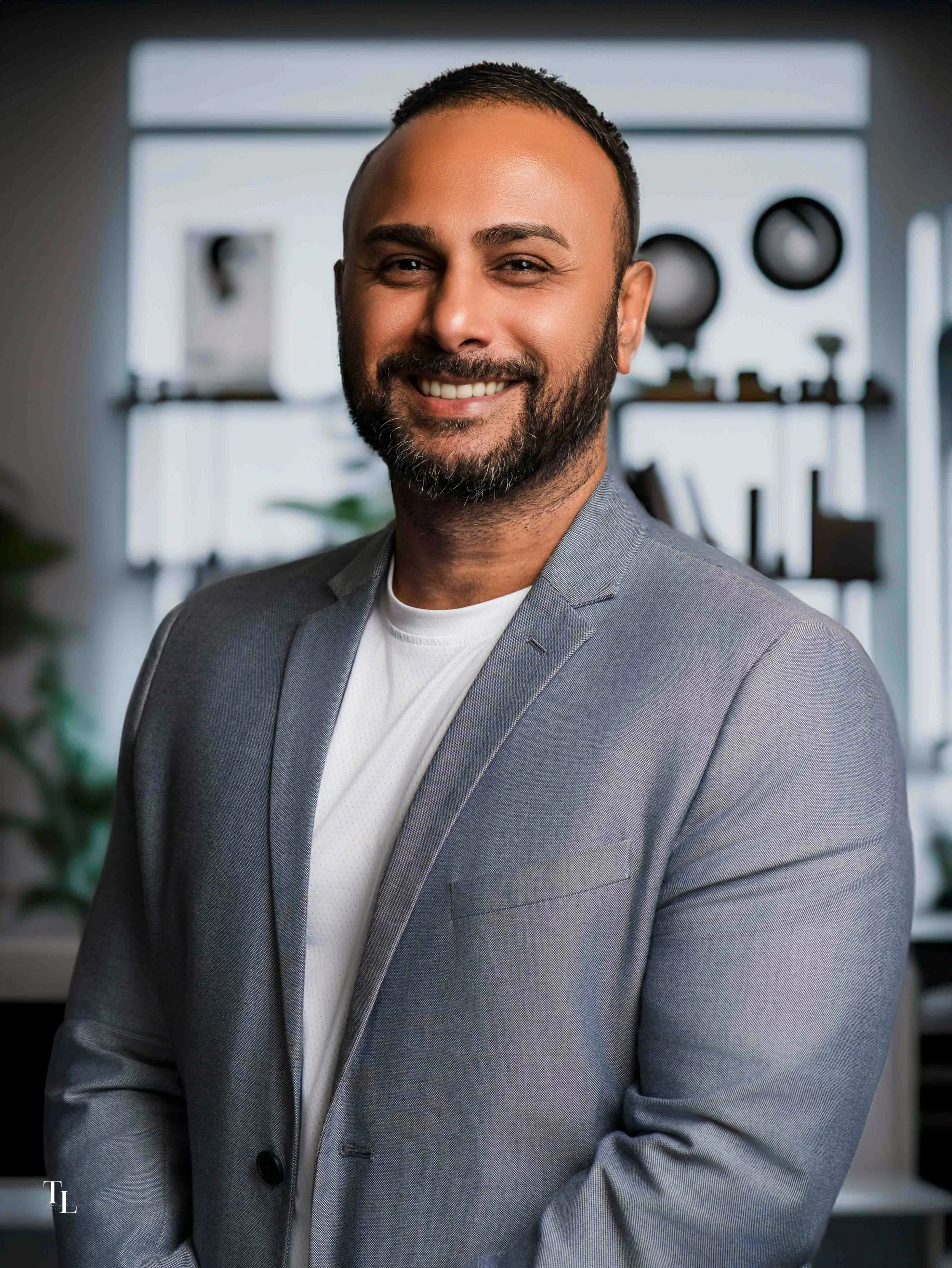 Smiling man in a gray blazer and white shirt, with a beard and short hair, in an office setting with a blurred background of shelves and speakers.