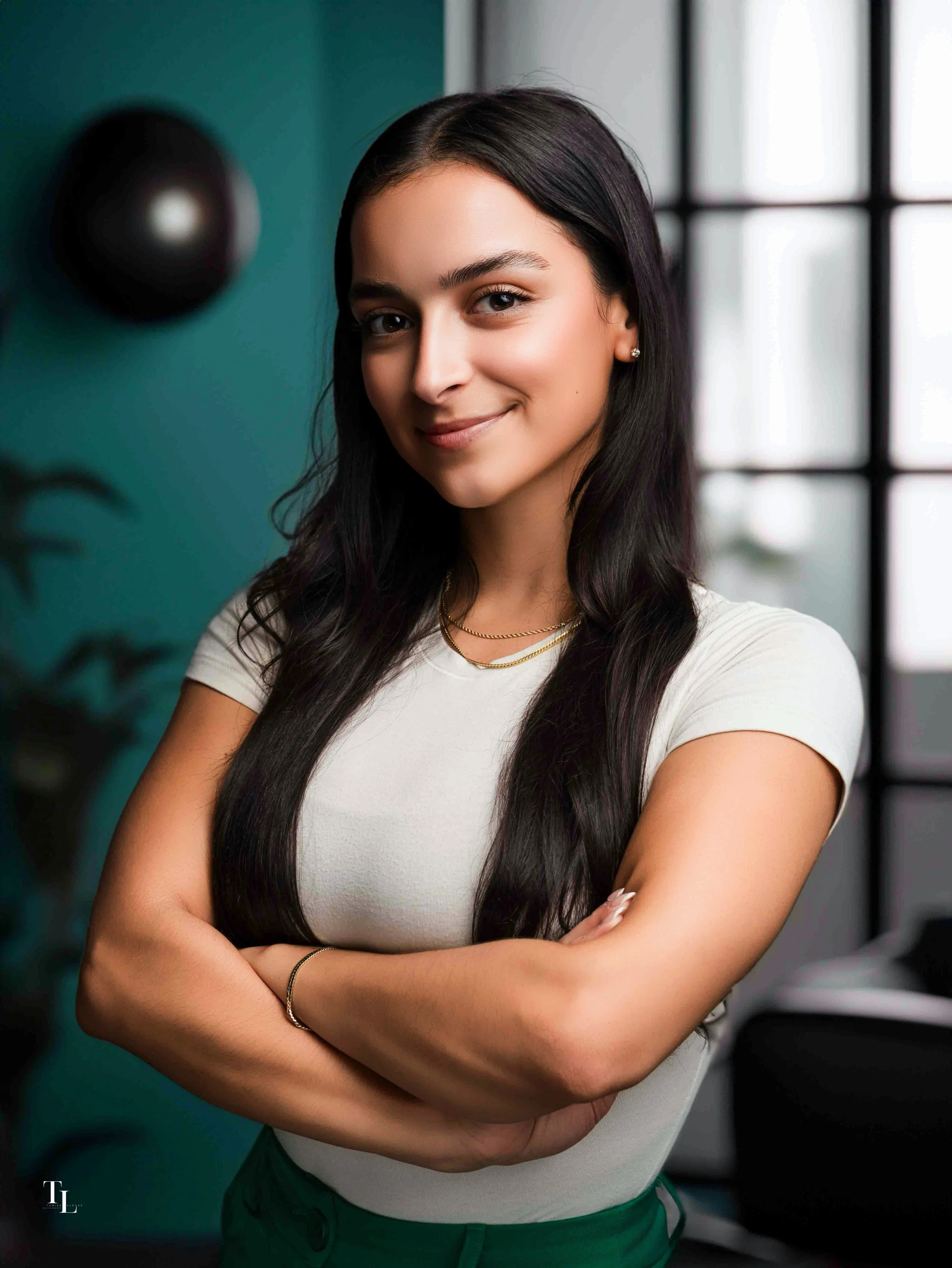 A young woman with long dark hair, wearing a white top and green pants, standing indoors near a window with a grid pattern, smiling gently with arms crossed, in a modern interior setting.