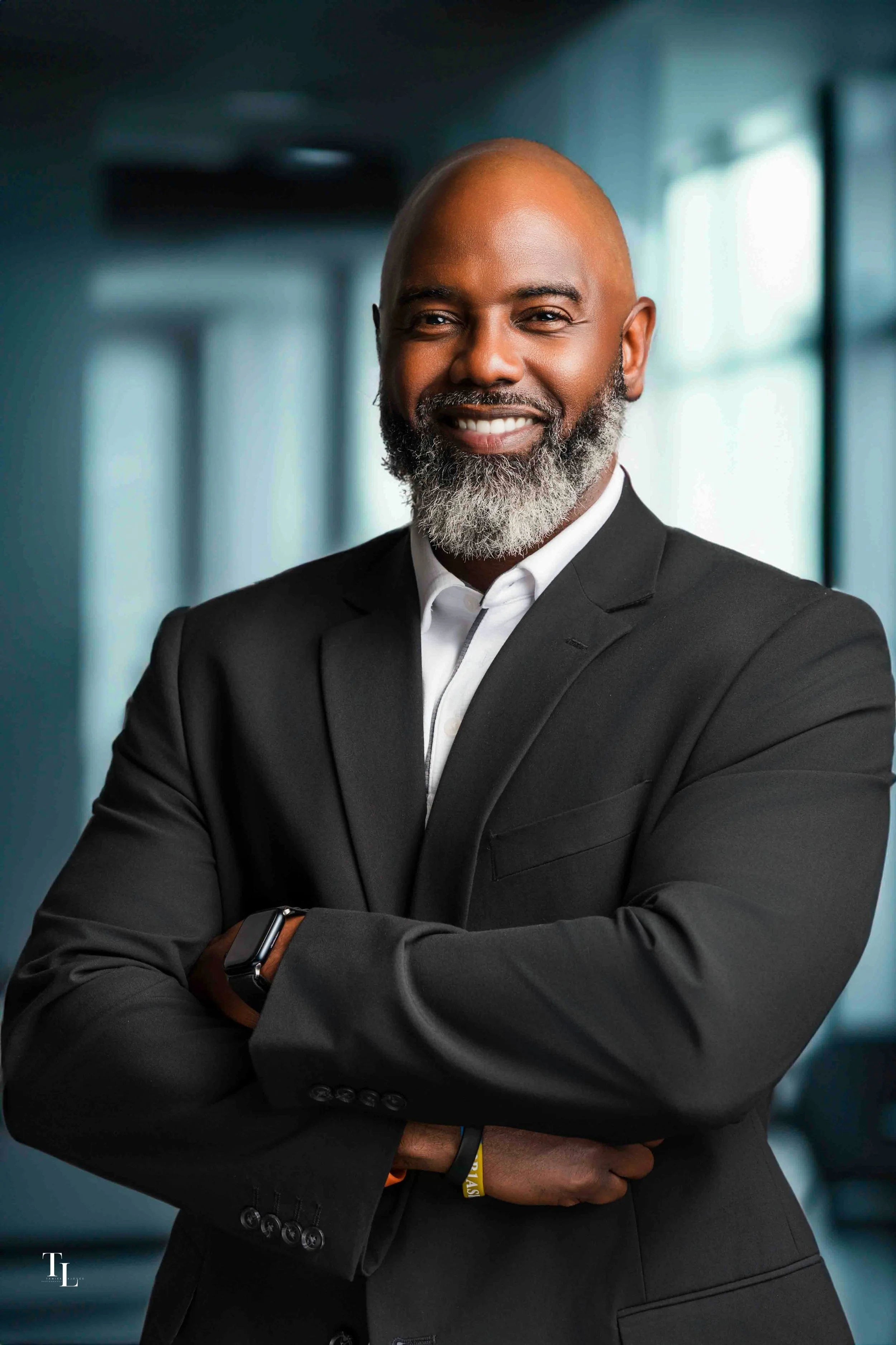 A confident African American man with a bald head and a gray beard, dressed in a black suit and white shirt, standing with arms crossed in a modern office or conference room with large windows and blurred background.