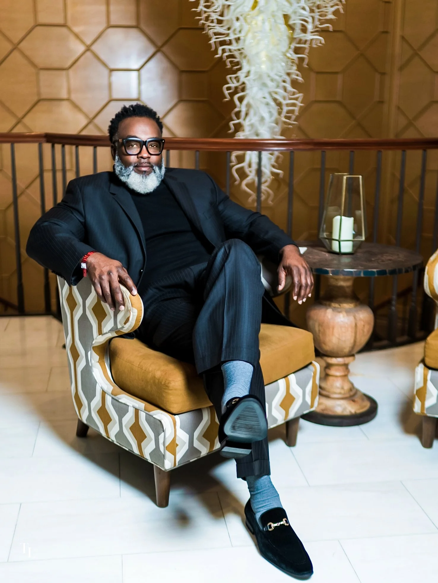 A man with a beard and glasses sitting on a patterned armchair in a room with wood-paneled wall and decorative sculpture behind him, next to a small round table with a lantern and candles, wearing a black suit and loafers.