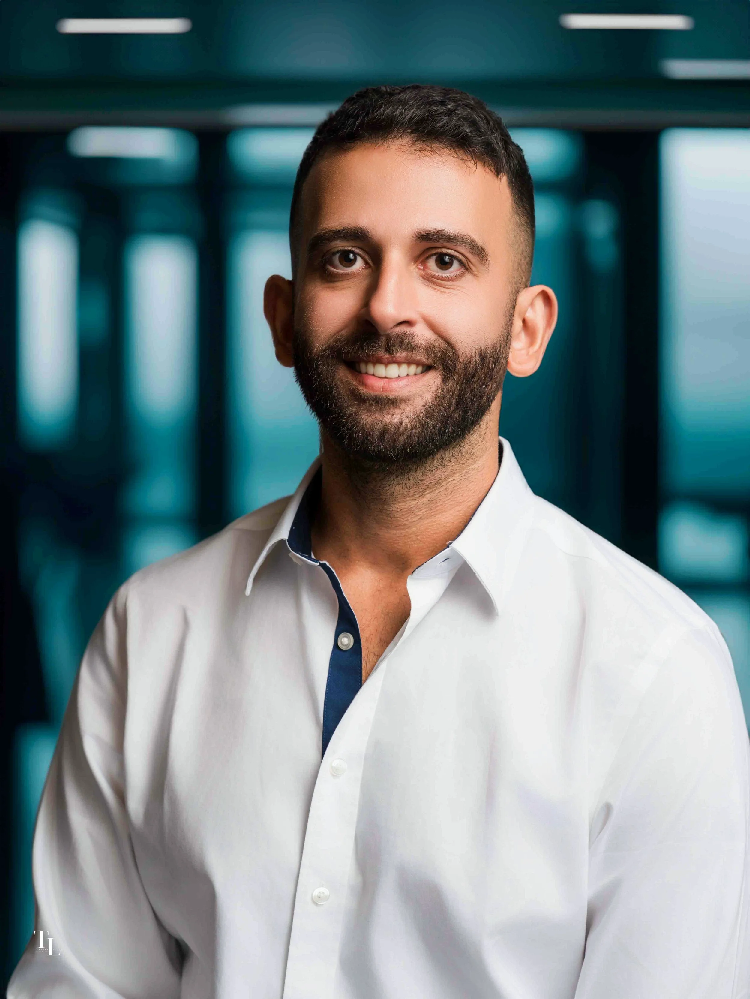 Headshot of a smiling man with short dark hair and a beard, wearing a white shirt, in an office or corporate environment.