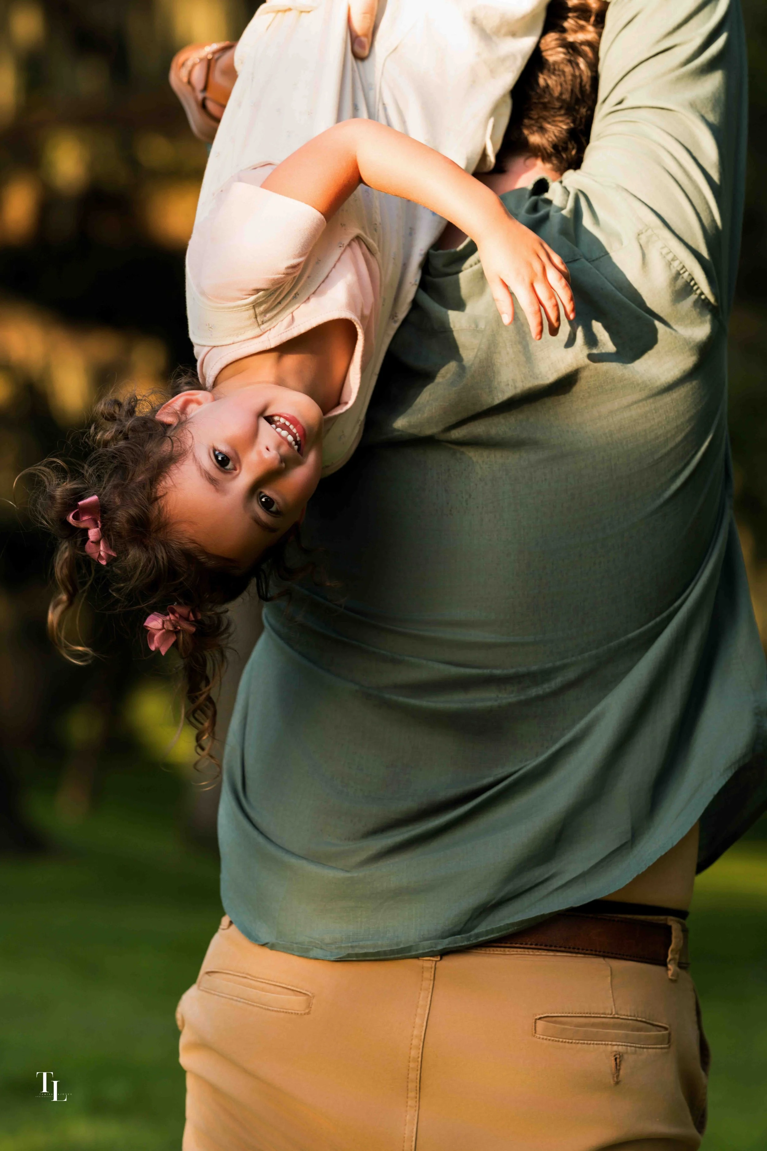 A man holding a smiling young girl upside down outdoors with trees and greenery in the background.