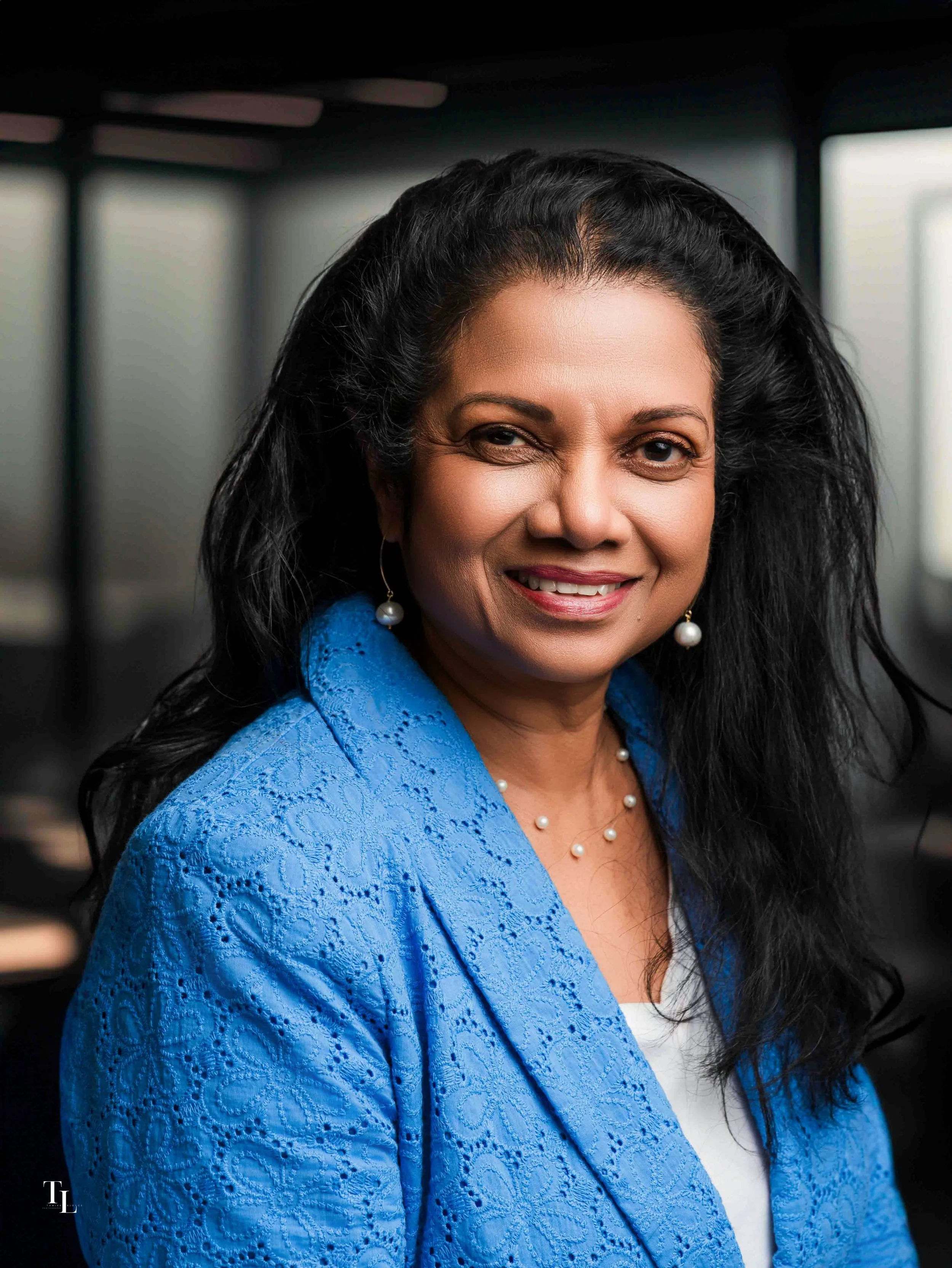 Close-up of a woman with black hair wearing a bright blue embroidered blazer, white top, pearl earrings, and necklace, smiling and winking. Background is dark and blurred.