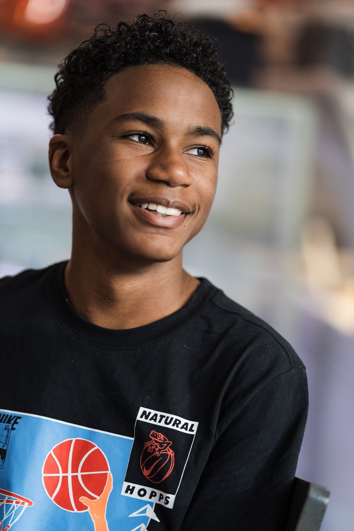 Young boy with curly hair smiling, wearing a black T-shirt with a basketball graphic and the words 'Natural Hops.'