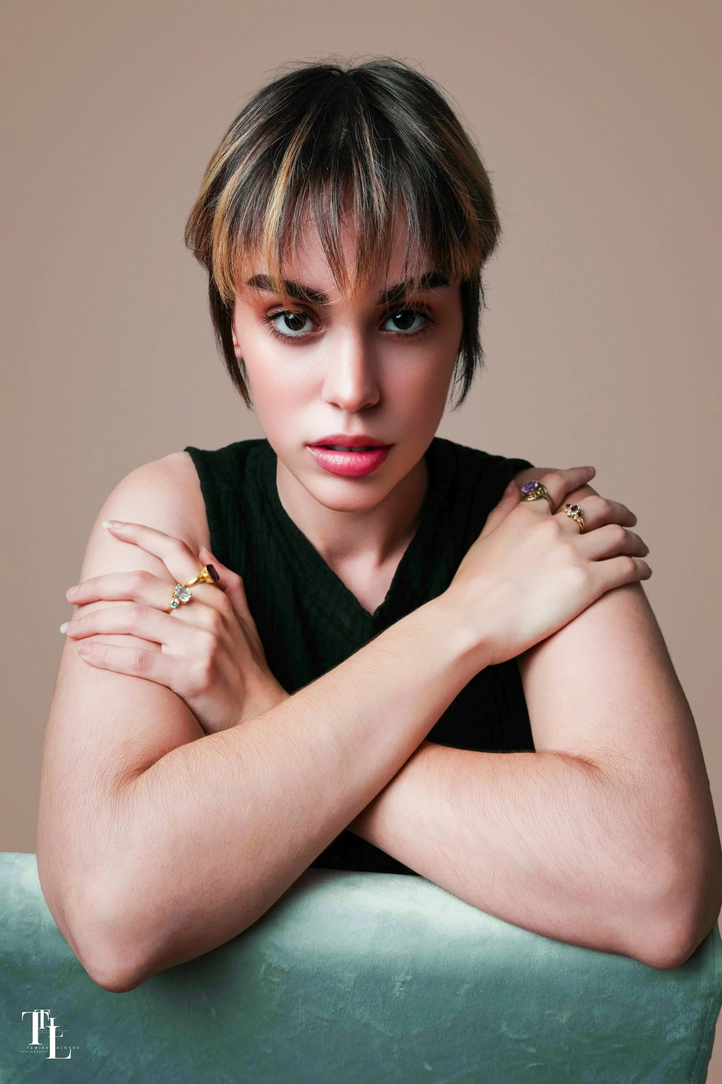 A young woman with short, tousled hair with blonde highlights, wearing a sleeveless black top, resting her arms crossed on a green-greenish surface, with a neutral beige background.