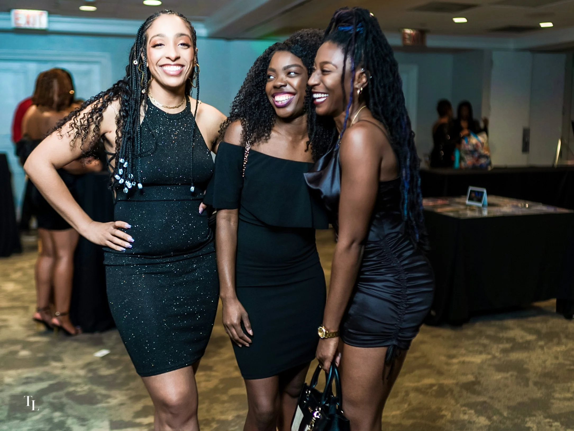 Three women smiling at a social event, dressed in black attire, standing close together.