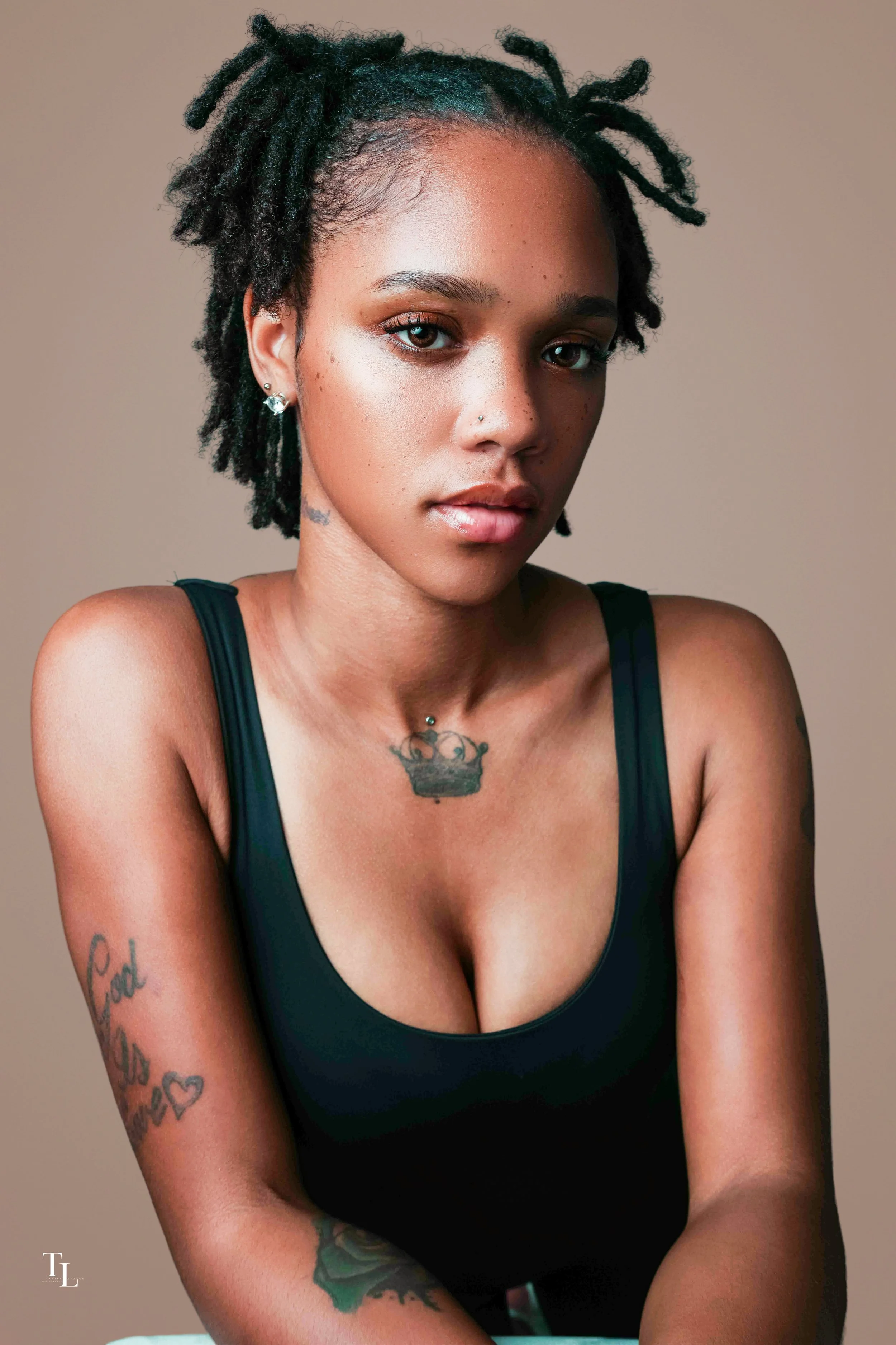 A young woman with short, curly, dark hair and light brown eyes looking directly at the camera. She has a nose piercing, diamond earrings, and tattoos on her neck and arms. She is wearing a black tank top and a neutral beige background.